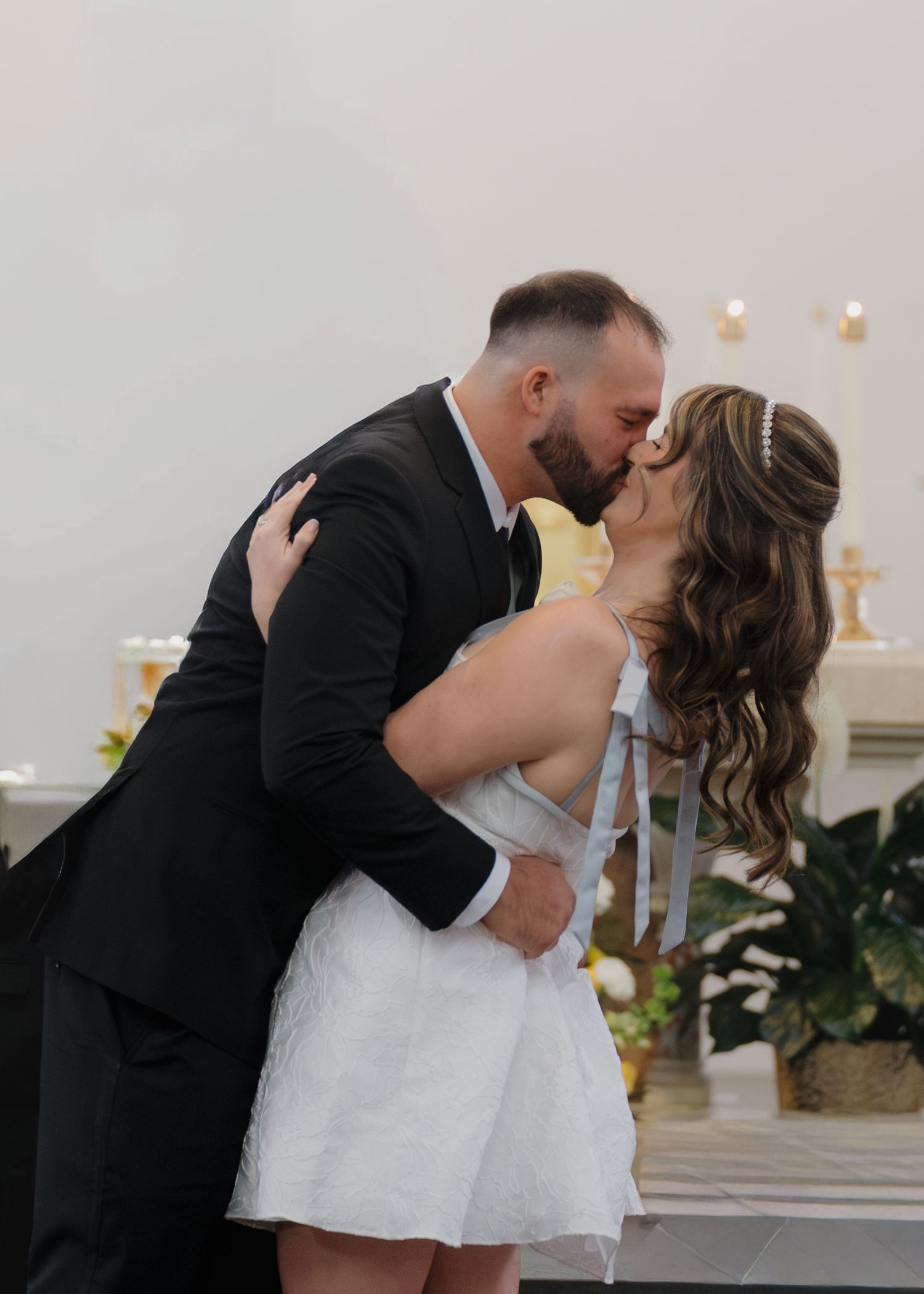 Bride and groom share first kiss at church altar, bride in short white dress with ribbon detail.
