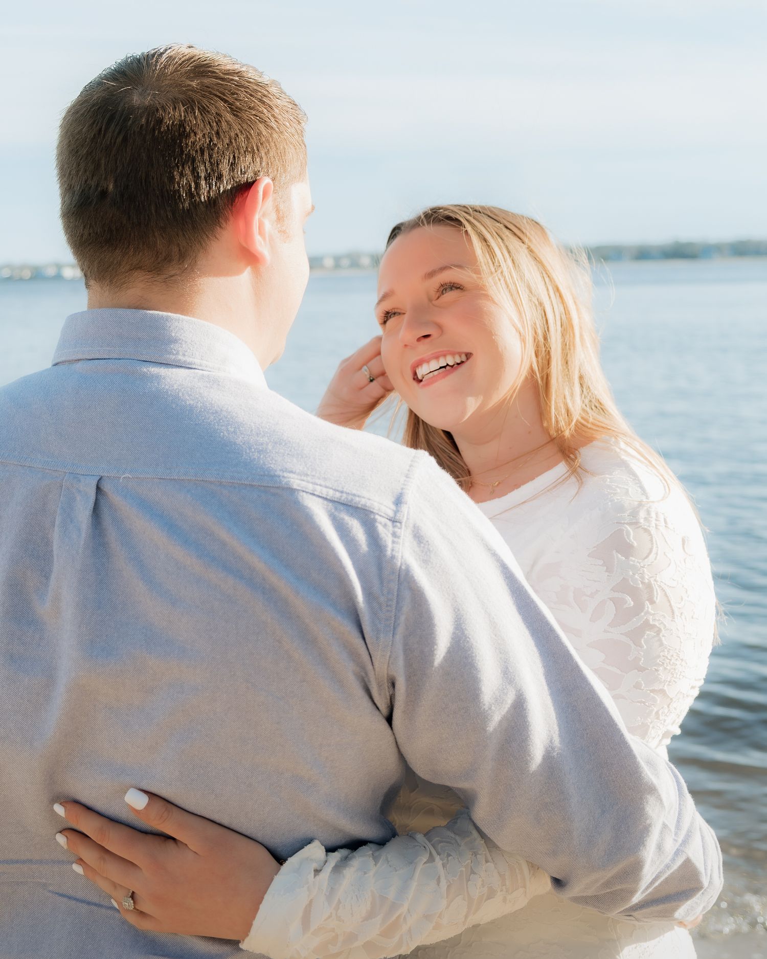 Couple embracing and smiling at each other near a waterfront on a sunny day, woman wearing white lace top.