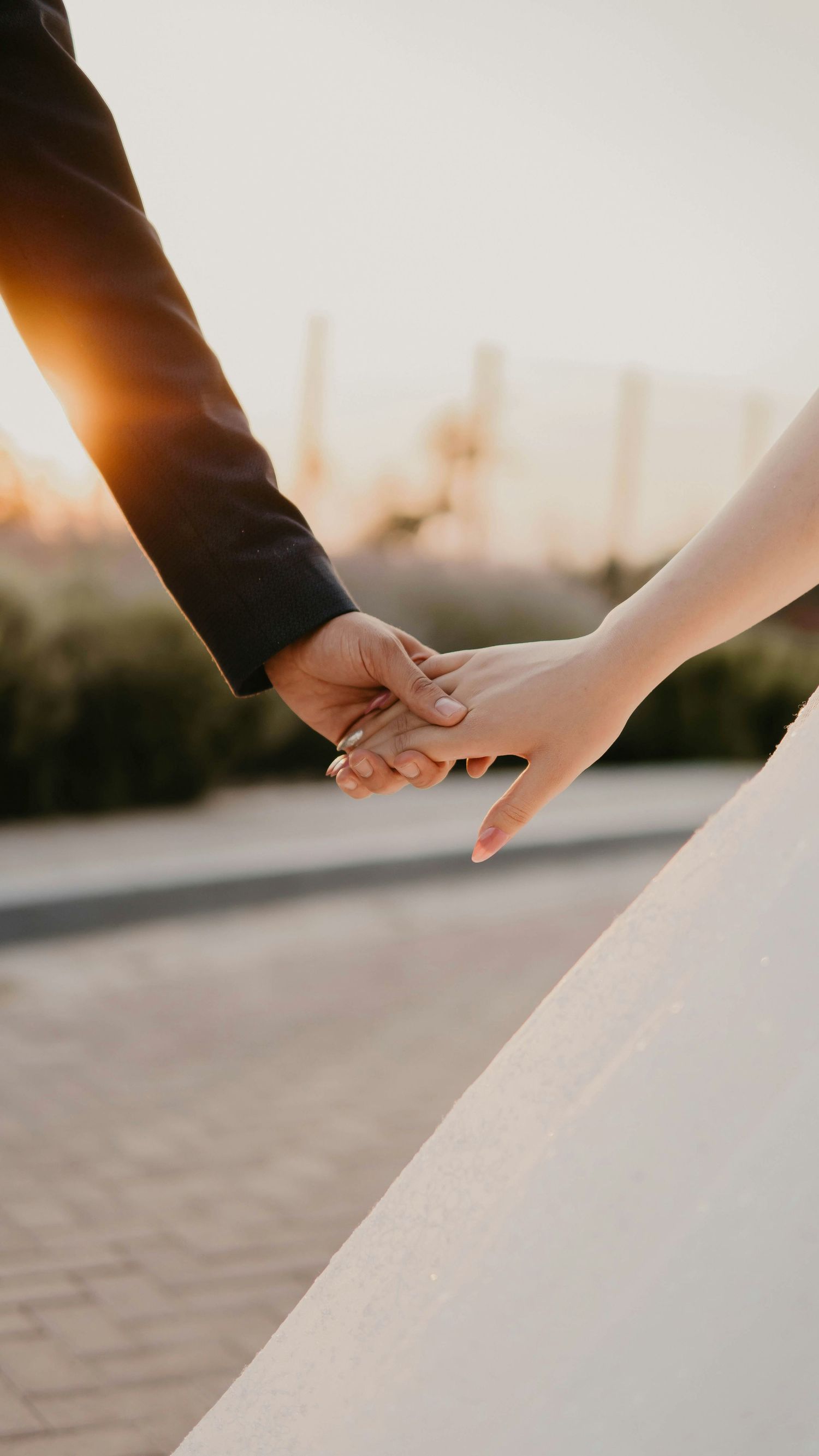 A bride and groom holding hands at sunset, with her white wedding dress and his dark suit visible.