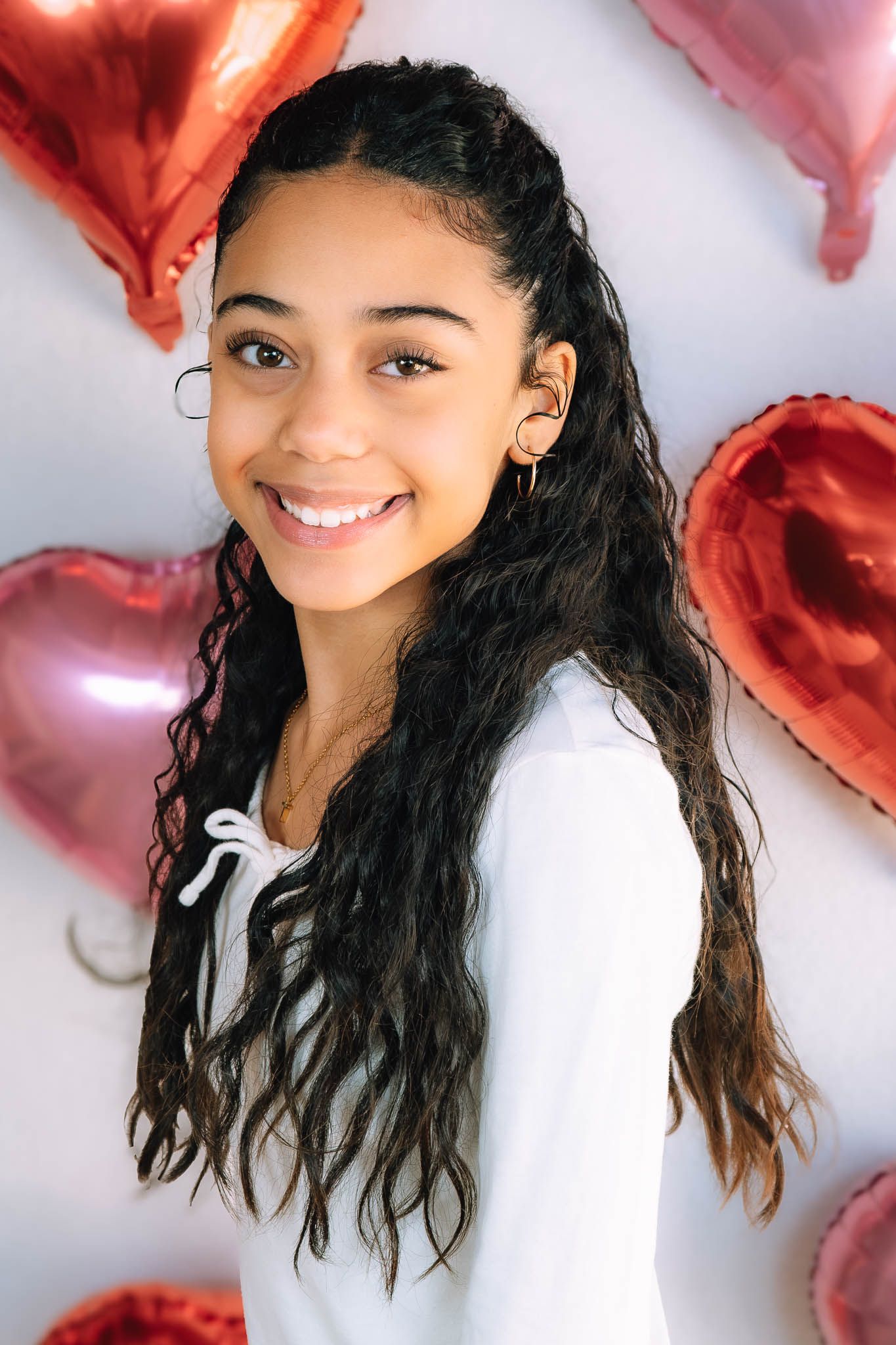 A smiling teenage girl with long curly braids poses in front of red and pink heart-shaped balloons.