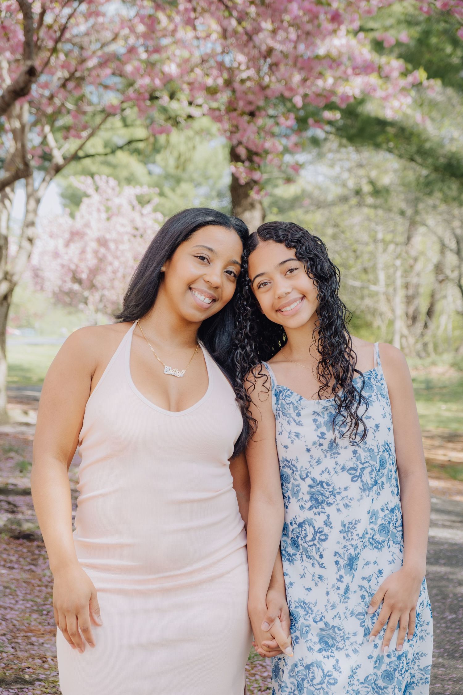 Two young women smiling outdoors in spring dresses under blooming cherry blossom trees in a park.