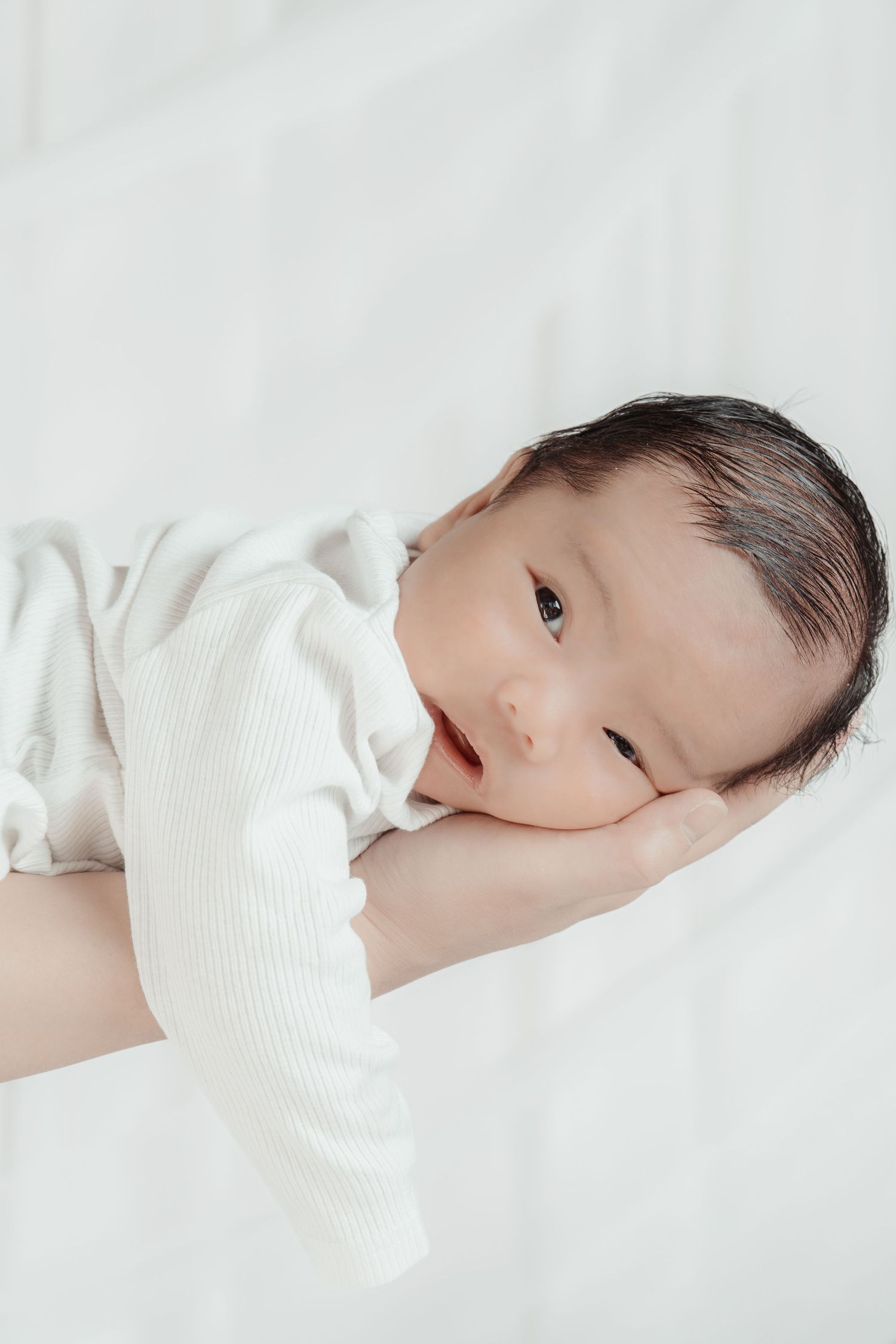 Newborn baby in white onesie resting peacefully on an adult's hand against a soft white background.