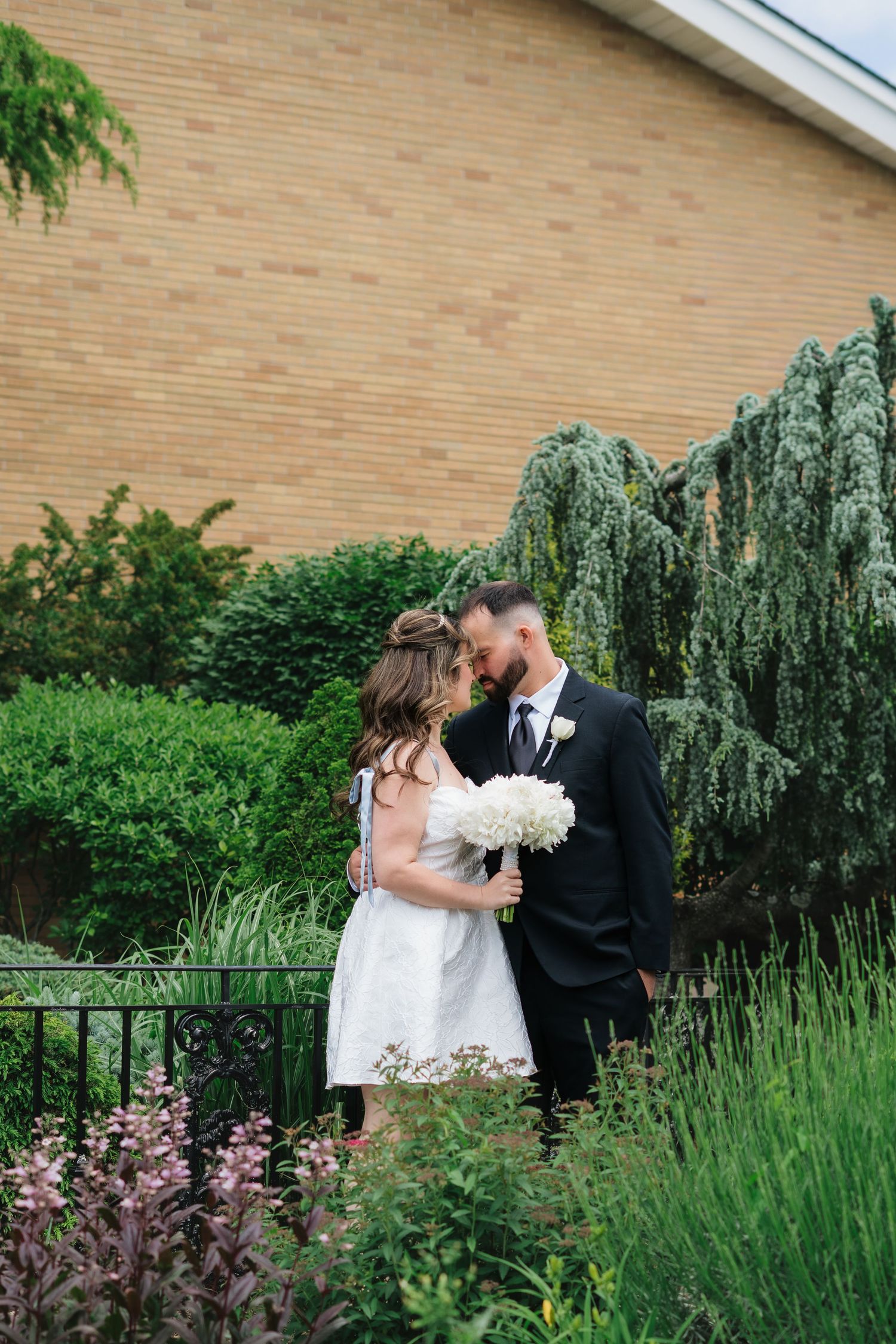 Bride and groom share intimate moment in lush garden, bride in white dress holding white bouquet outdoors.