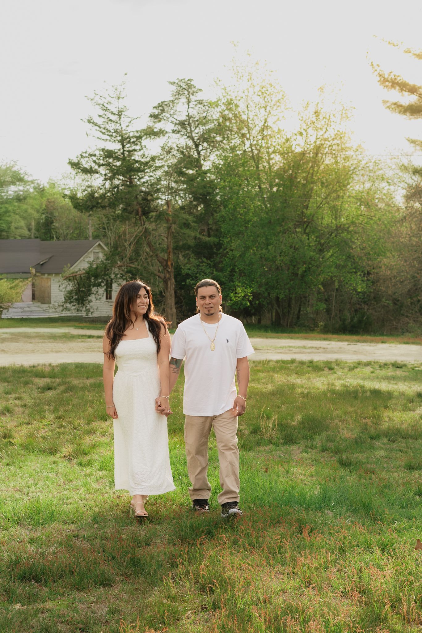 Couple holding hands in a green field, woman in white dress, man in white shirt and khaki pants, trees and house in background.
