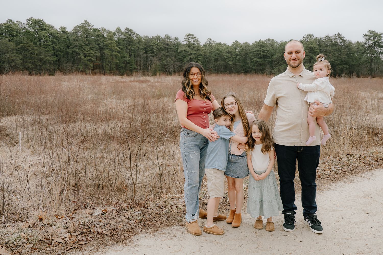 A smiling family of six stands together on a sandy trail with a pine forest backdrop.