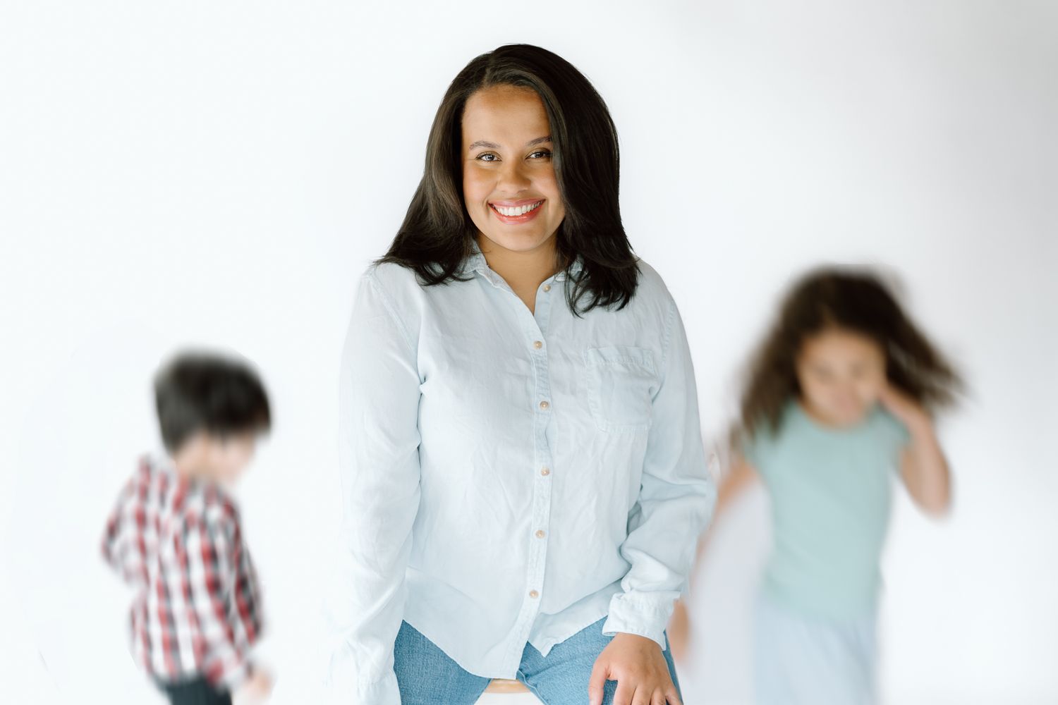 Smiling woman in white shirt poses confidently while two children play in the blurred background.