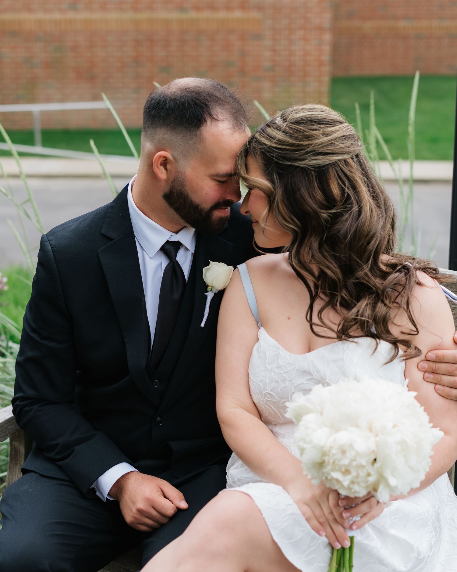 A bride and groom share an intimate moment, touching foreheads while seated outdoors, she holds white hydrangeas.