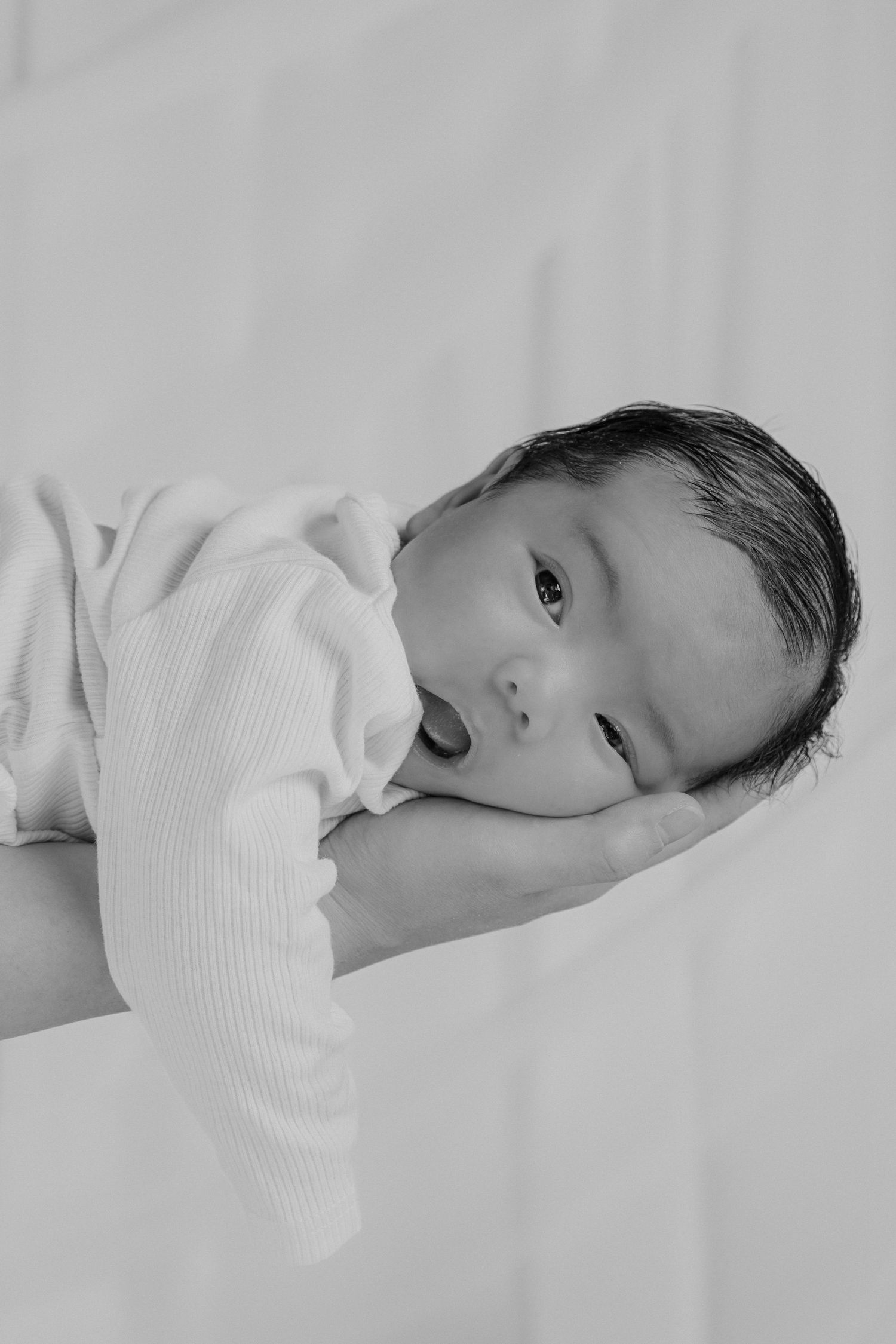 Black and white photo of a newborn baby cradled in an adult's hand, wearing a white onesie.