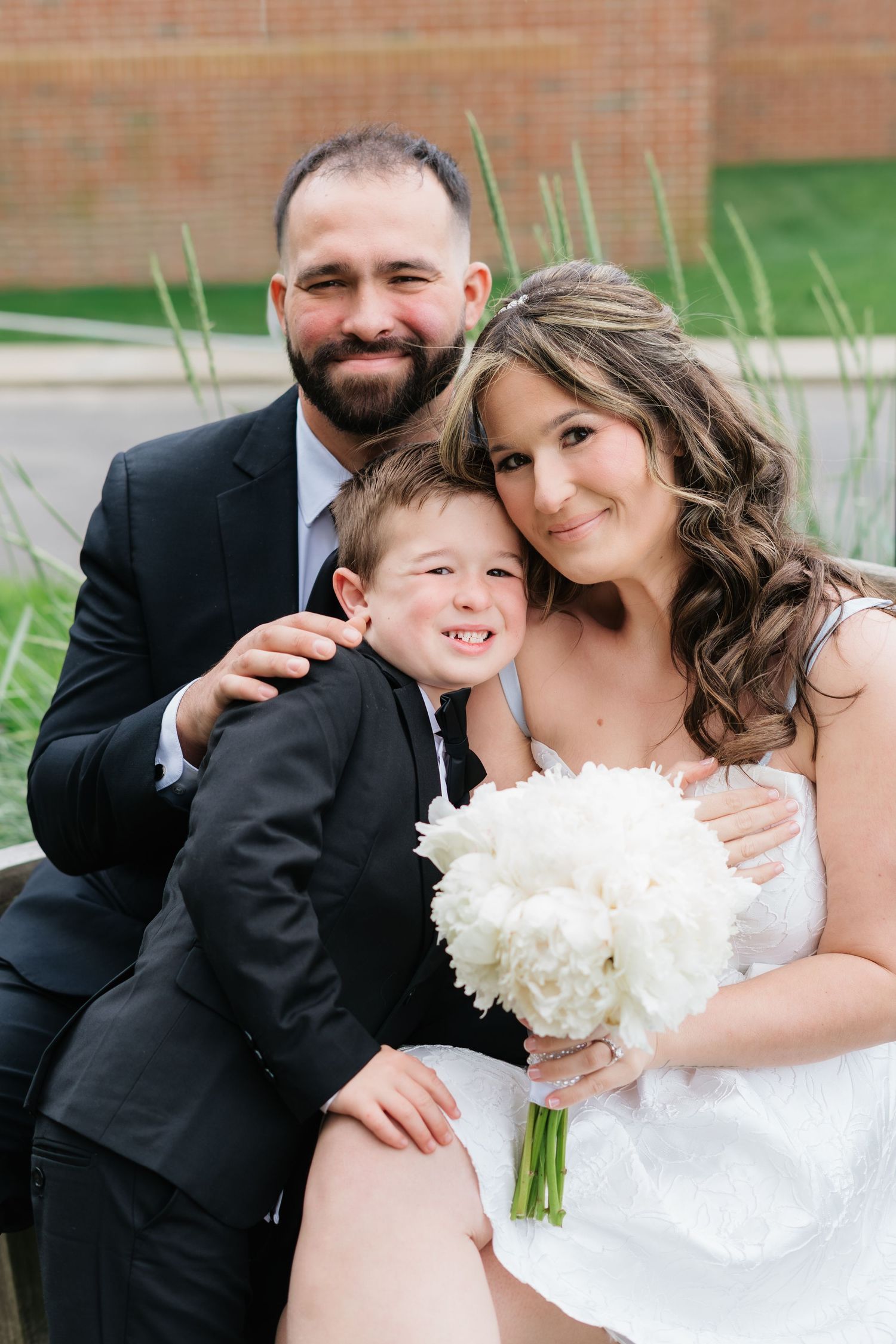 Smiling bride and groom sit with young boy in suit, bride holding white peony bouquet outdoors.