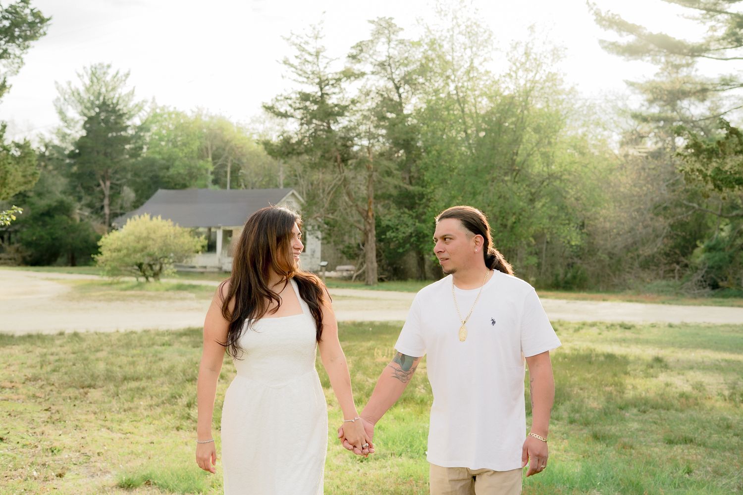 Couple walking hand in hand through a sunny green field, woman in white dress and man in white shirt smiling at each other.