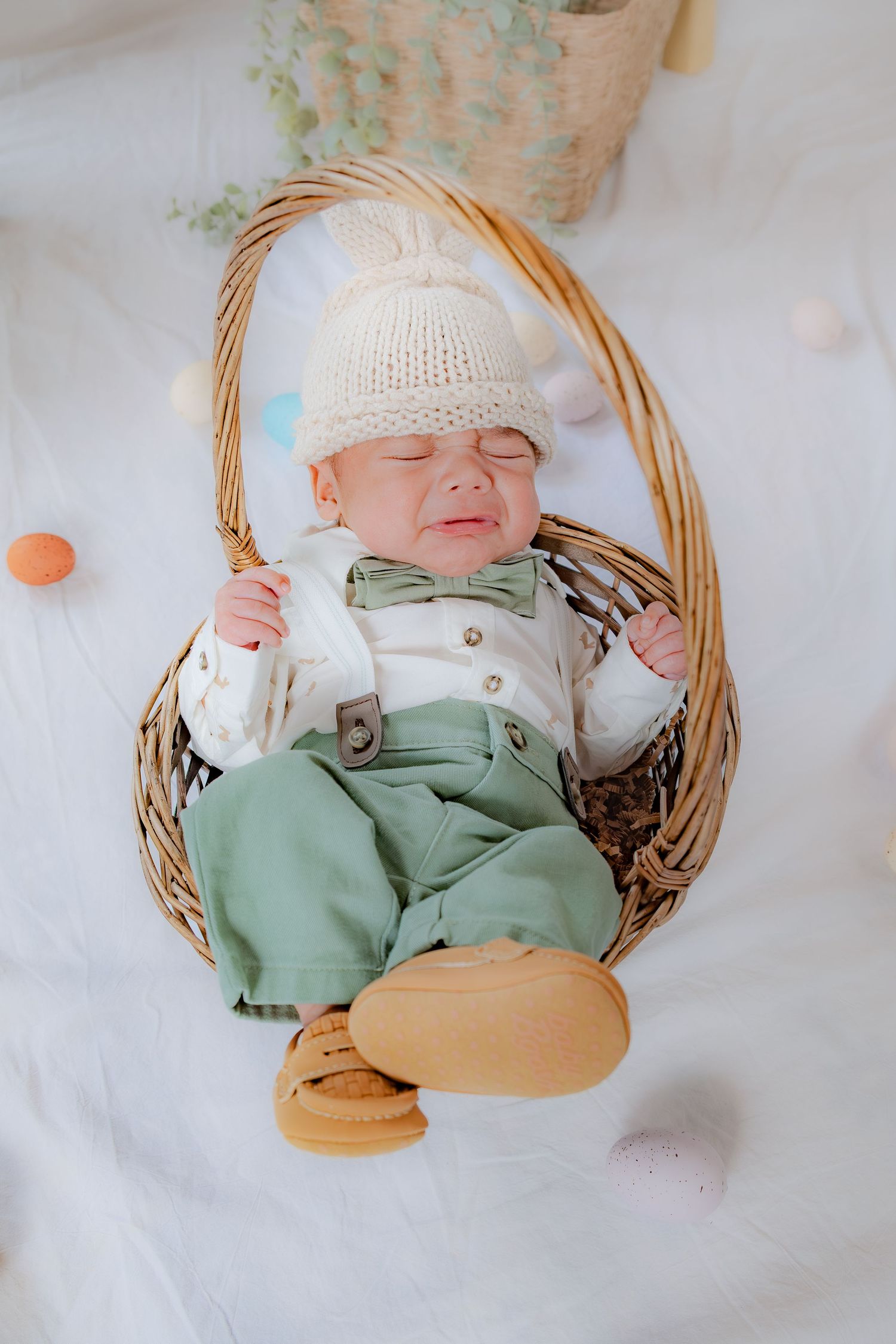 A newborn baby in green pants and a cream knit hat sleeps in a wicker Easter basket.