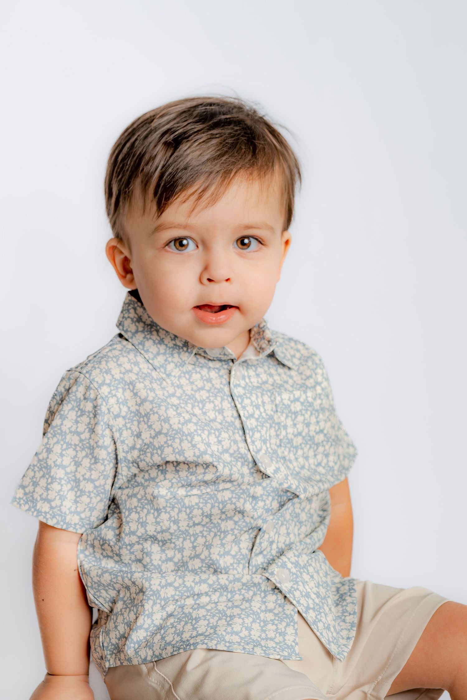 A cute toddler boy in a floral blue shirt and tan shorts sits against a white background.
