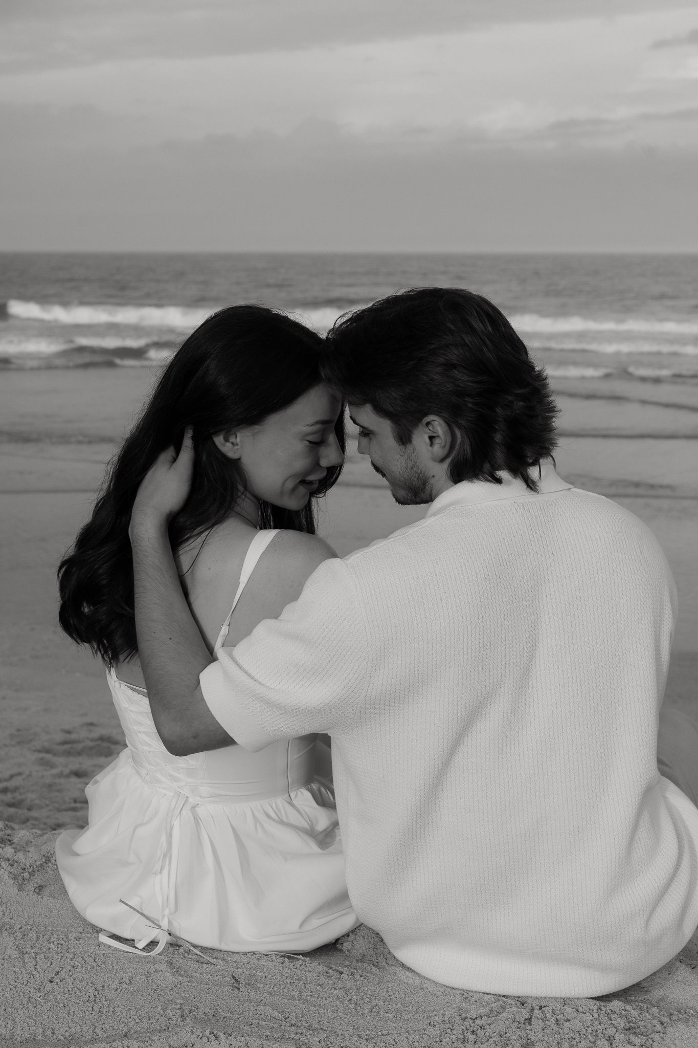 Black and white photo of a romantic couple sitting close together on a beach, foreheads touching tenderly.
