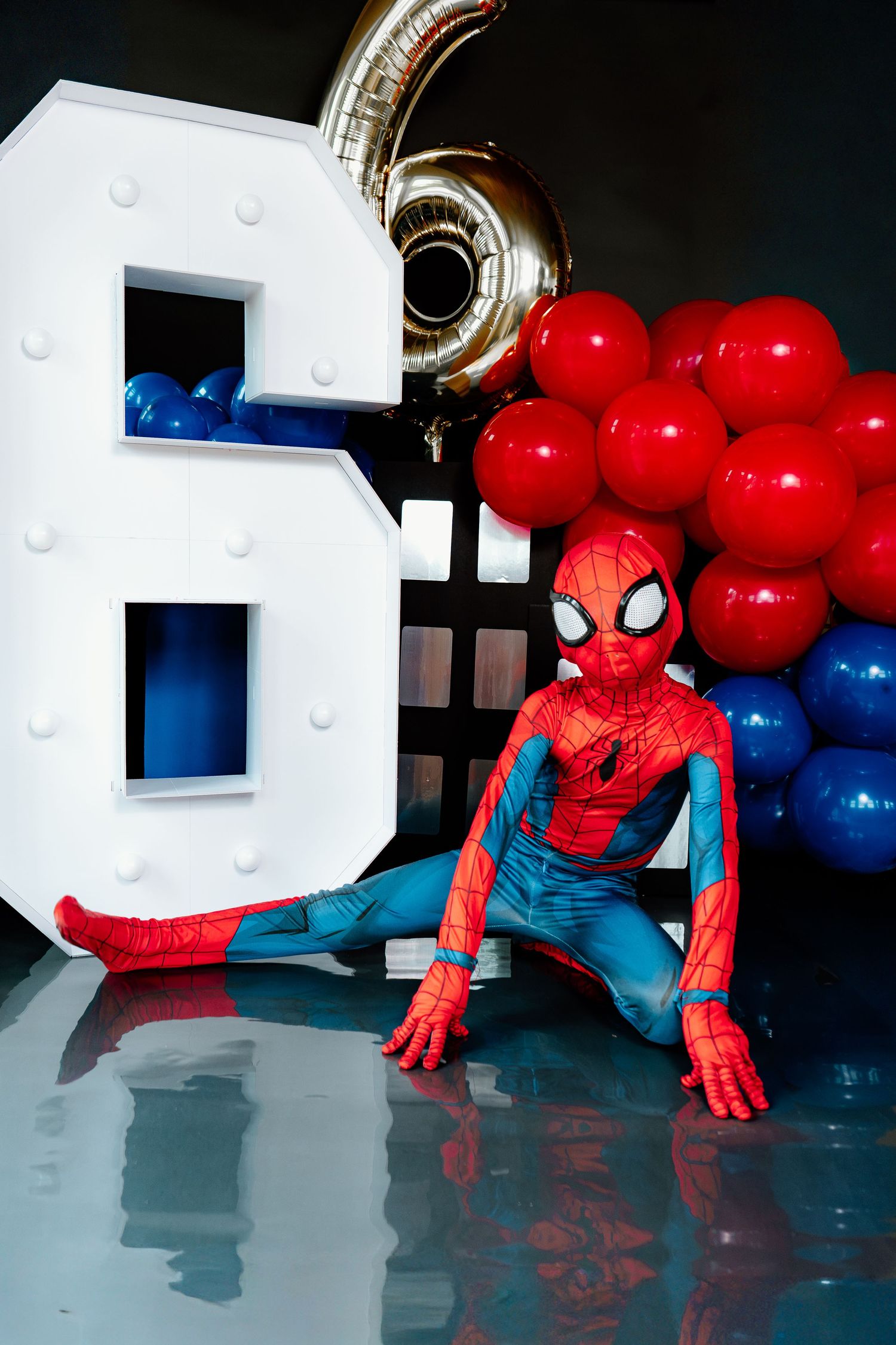 Child in Spiderman costume sitting by illuminated number 6 with red and blue balloons at birthday party.