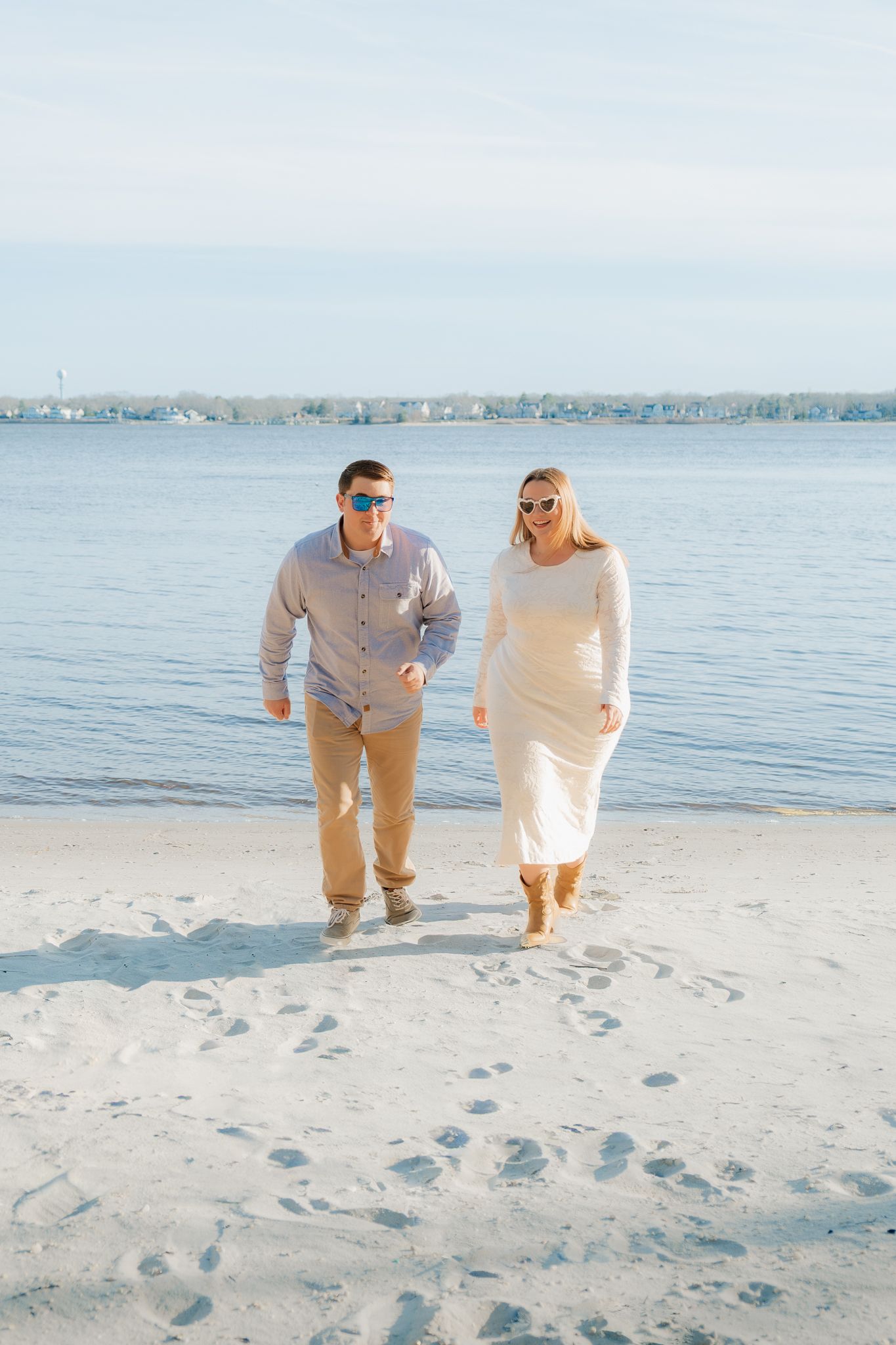 Couple in sunglasses walking hand in hand on a sandy beach near calm blue water on a sunny day.