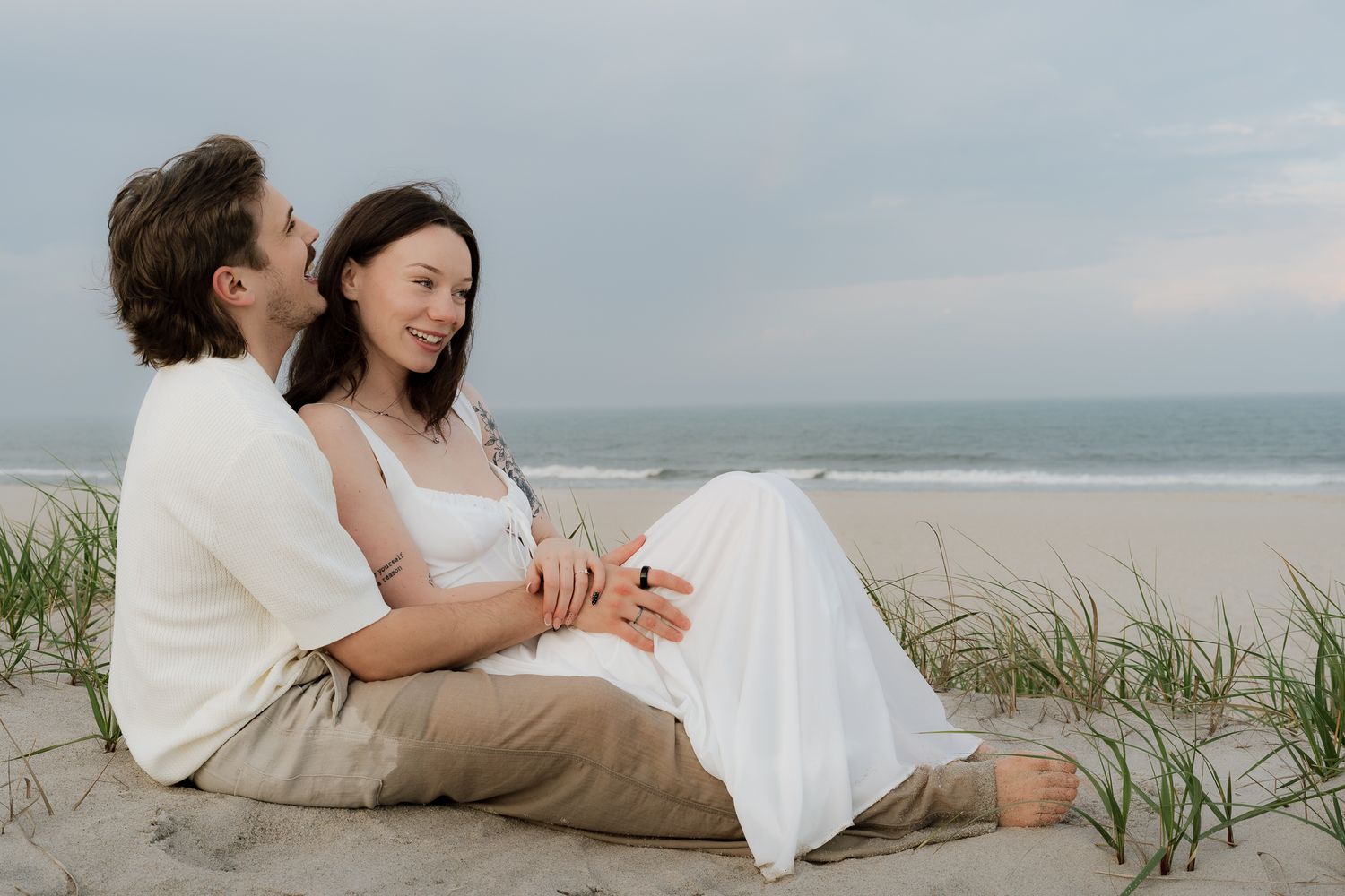 A couple sits together on a sandy beach, smiling and holding hands with ocean waves in the background.