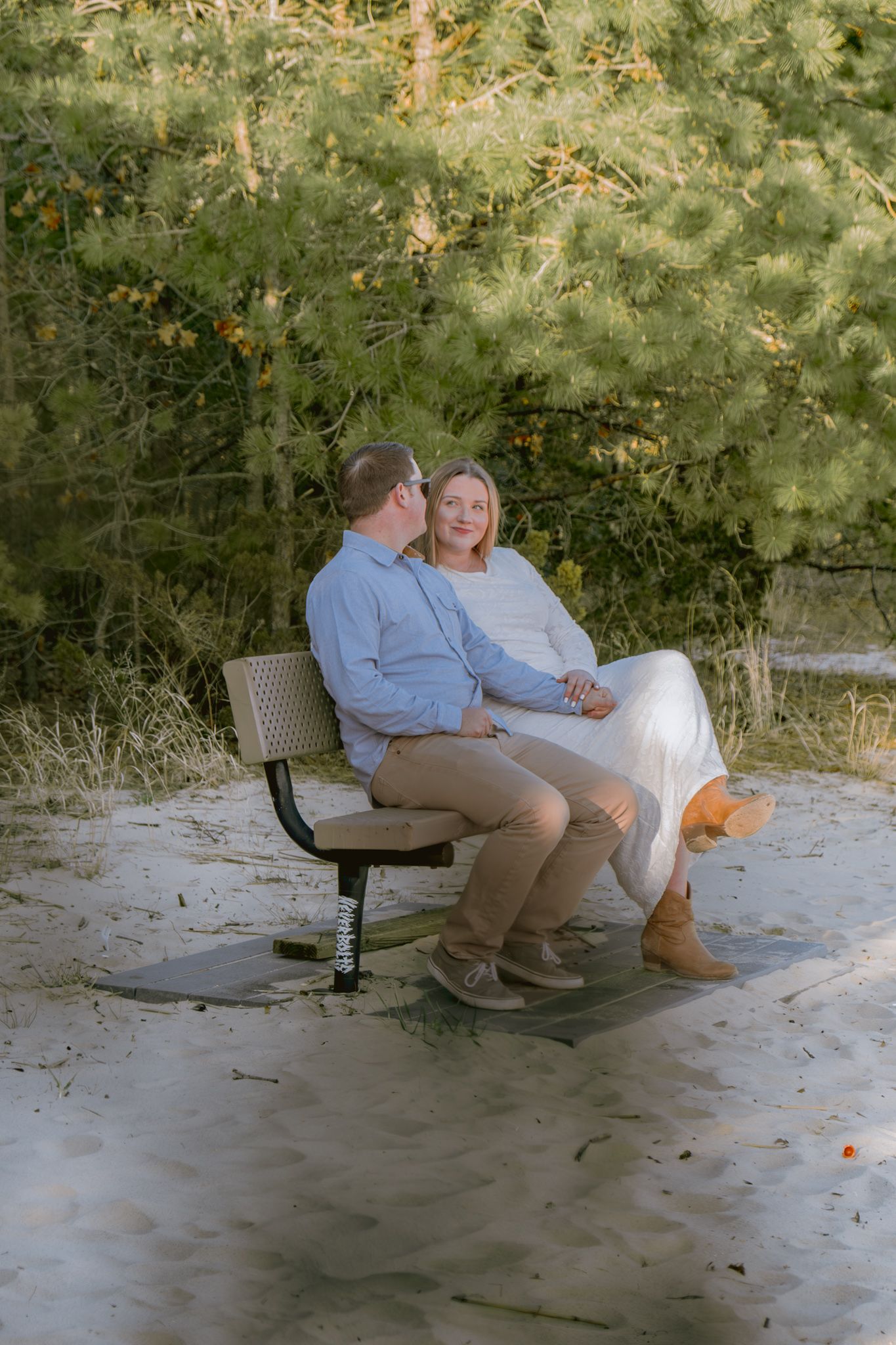 Engaged couple sitting together on a bench surrounded by sandy ground and lush green pine trees.