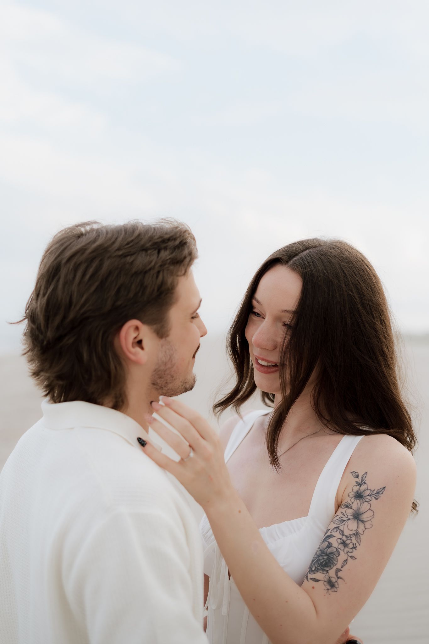 Smiling couple in white outfits share a tender moment on a beach, woman with floral arm tattoo touches mans face.