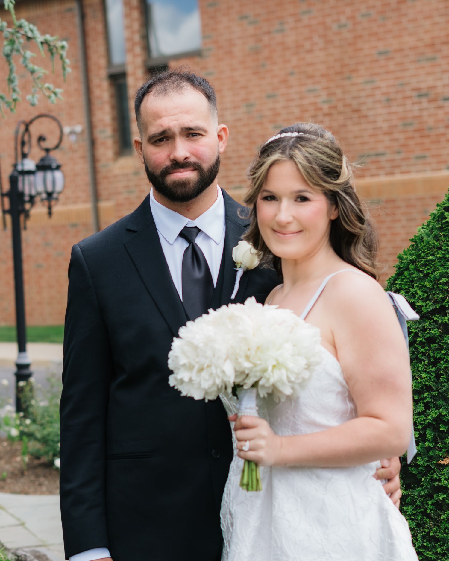 Bride and groom posing together outdoors on their wedding day, bride holding white floral bouquet.