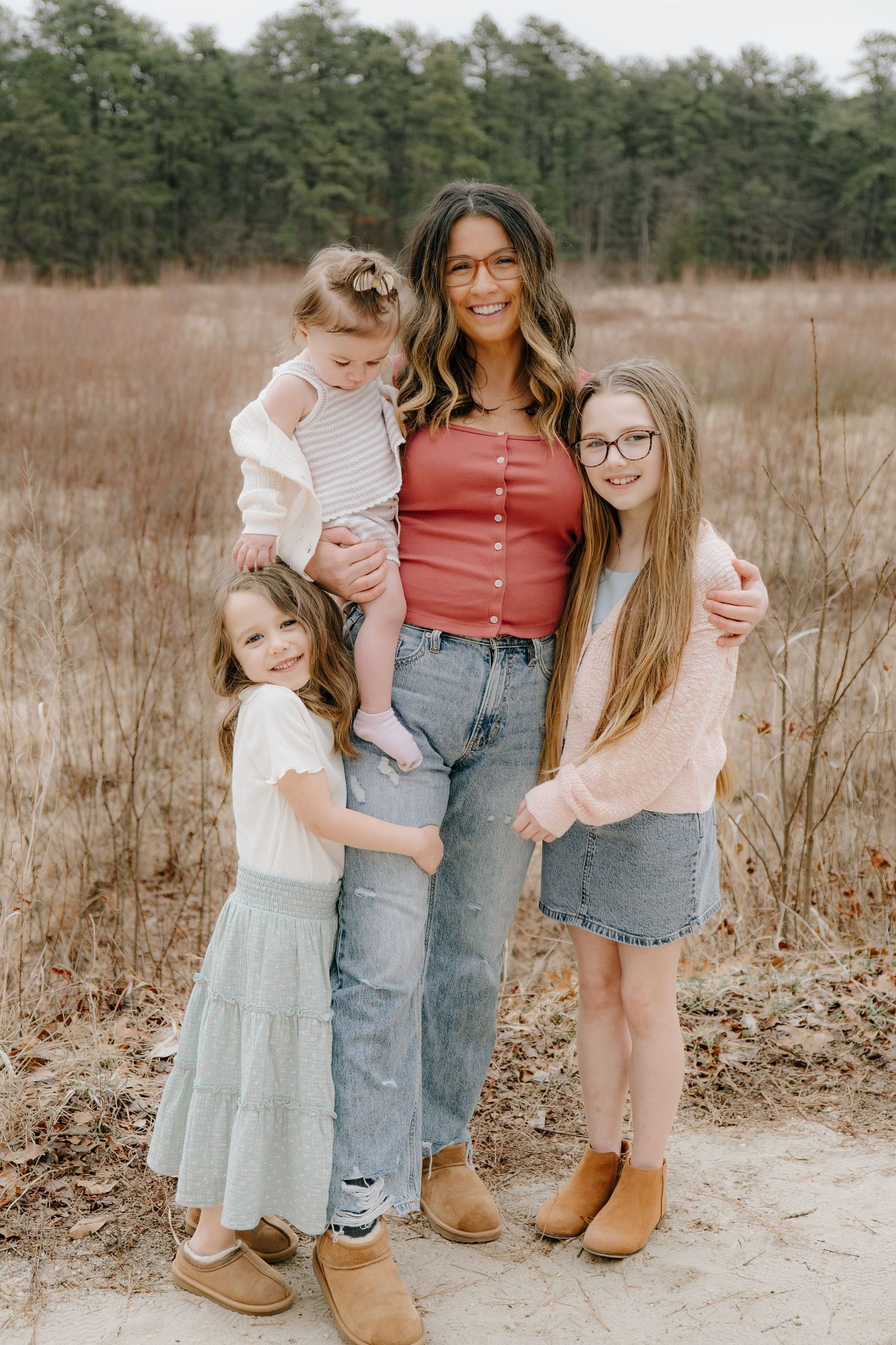 A woman in a red top and jeans poses outdoors with three young girls in a field with pine trees.