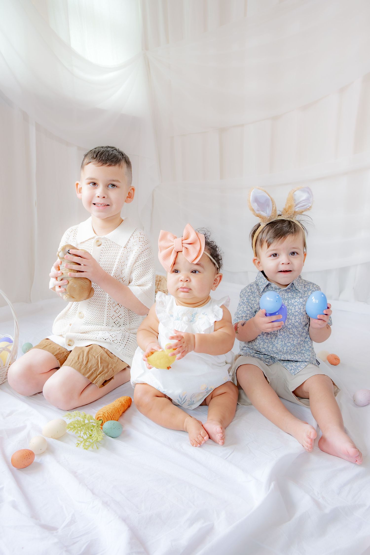 Three young children in Easter outfits sit together holding Easter eggs and toys against a white background.
