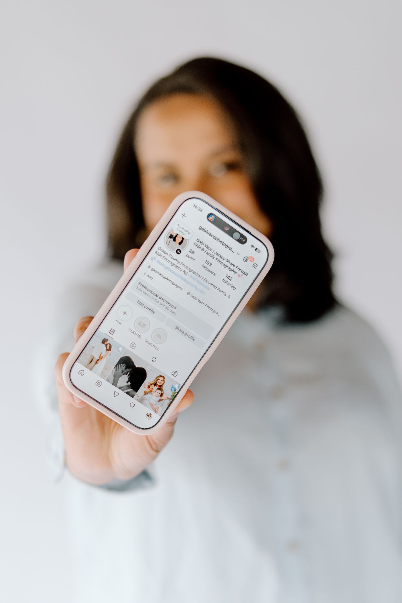 Woman holding smartphone displaying an Instagram profile page toward camera in a bright, minimalist setting.