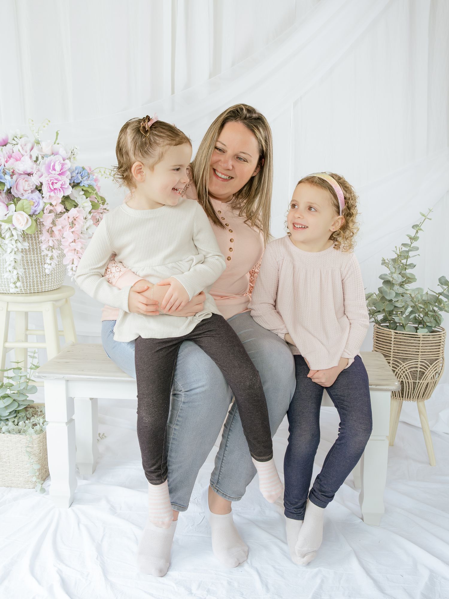 A smiling mother sits with her two young daughters in pink and cream outfits in a bright floral studio setting.