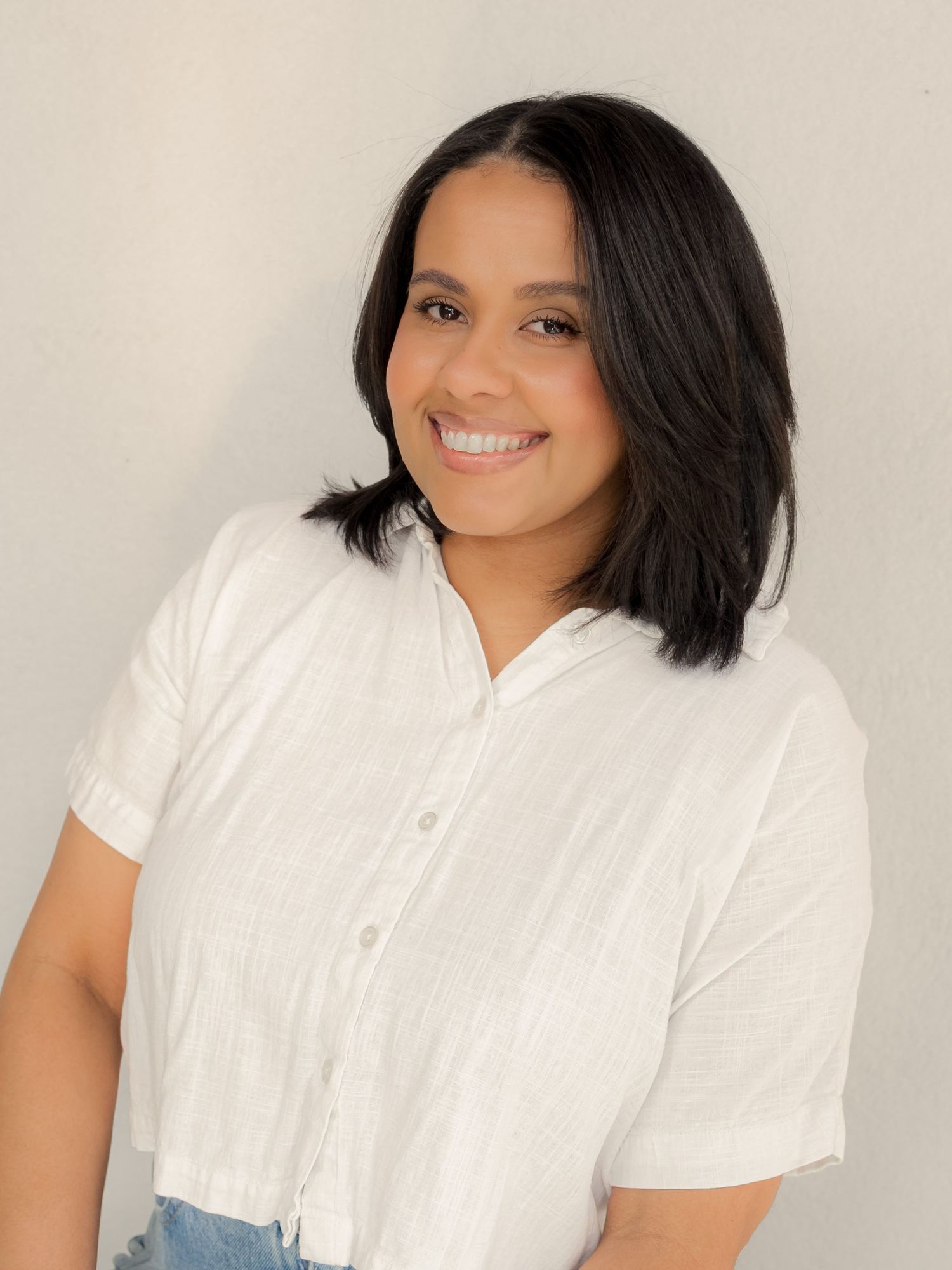 Smiling woman with dark bob haircut wearing white button-up shirt poses against light gray background.