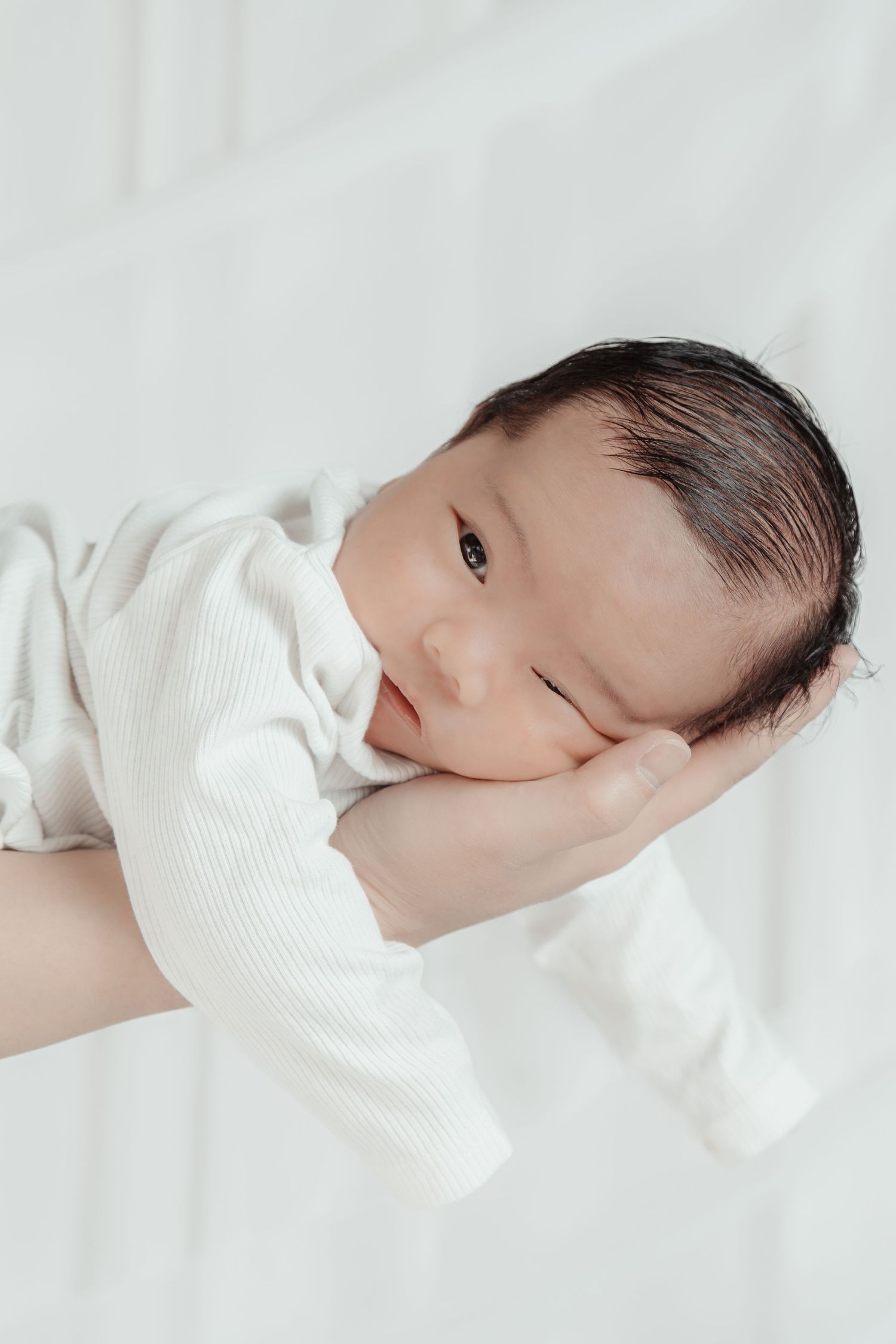 A newborn baby in a white onesie resting peacefully in a parent's hand against a soft white background.