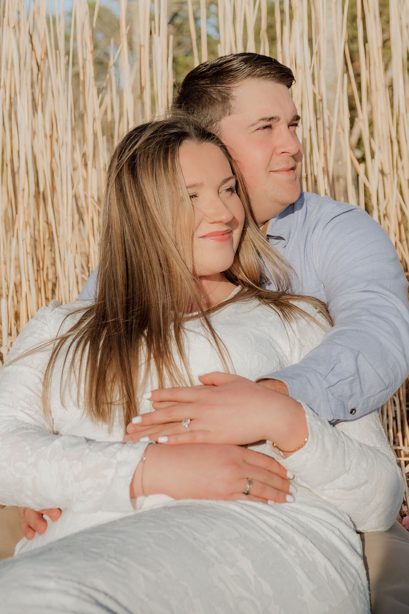 Couple embracing outdoors near tall golden reeds, woman wearing engagement ring in white outfit, man in blue shirt.