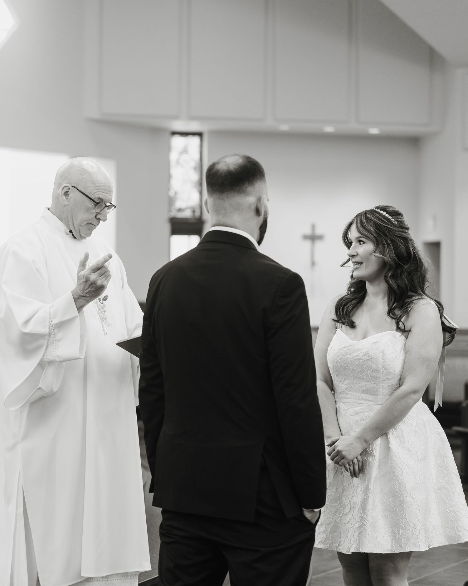 Black and white photo of a bride and groom exchanging vows with an officiant in a church ceremony.