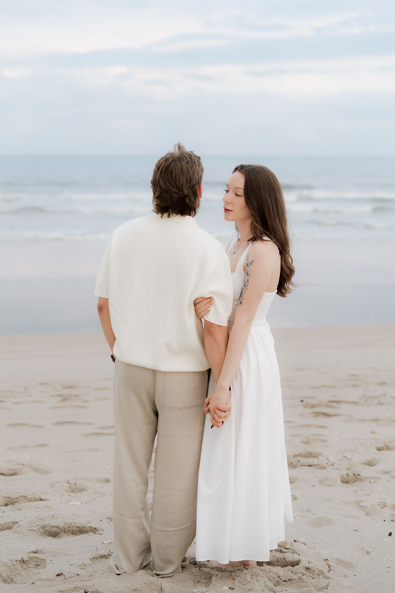A couple holds hands on a sandy beach, the woman in a white dress gazing at her partner with the ocean in the background.