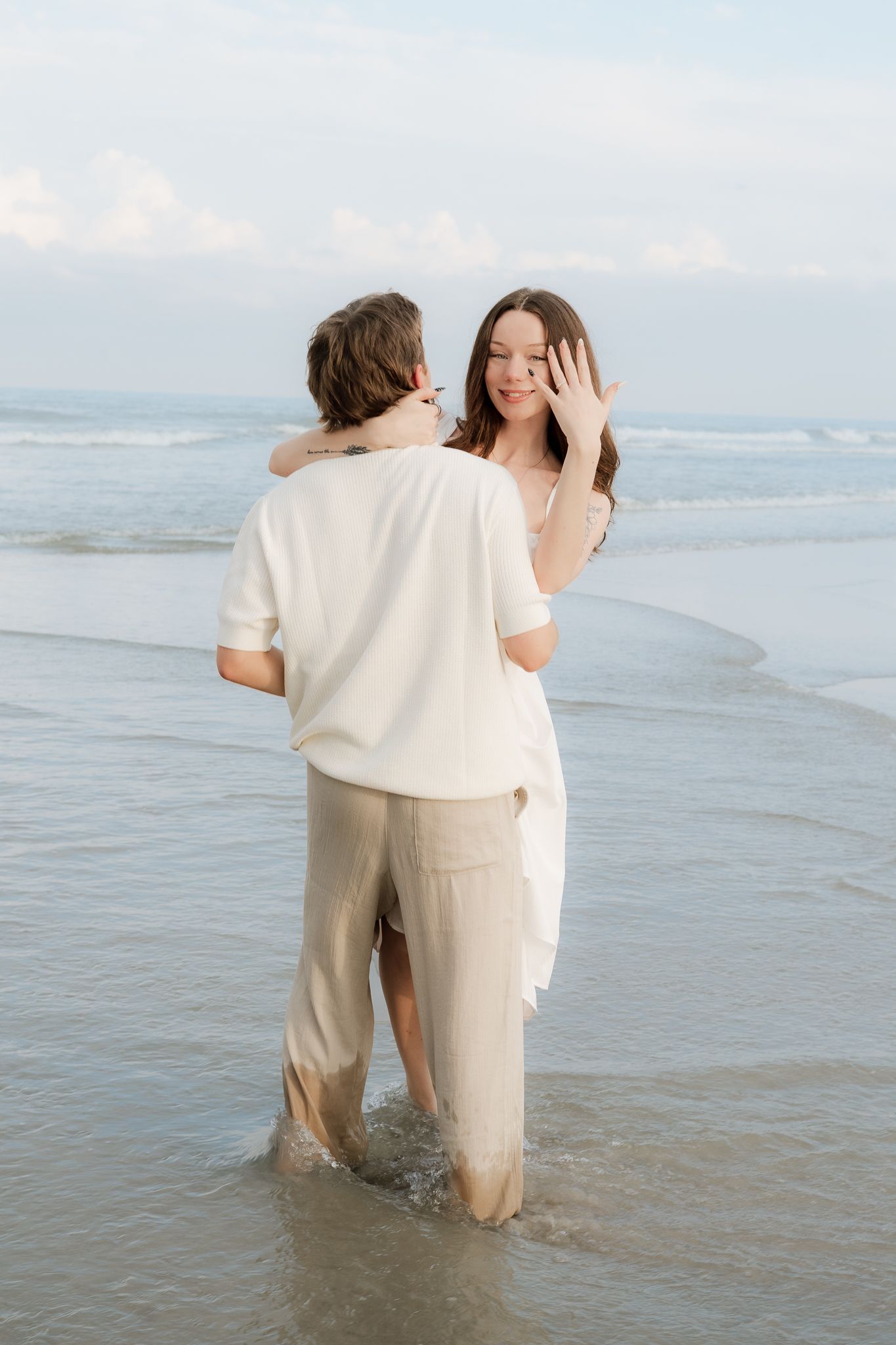 Couple standing in ocean waves, woman showing engagement ring while embracing man in white shirts on beach.