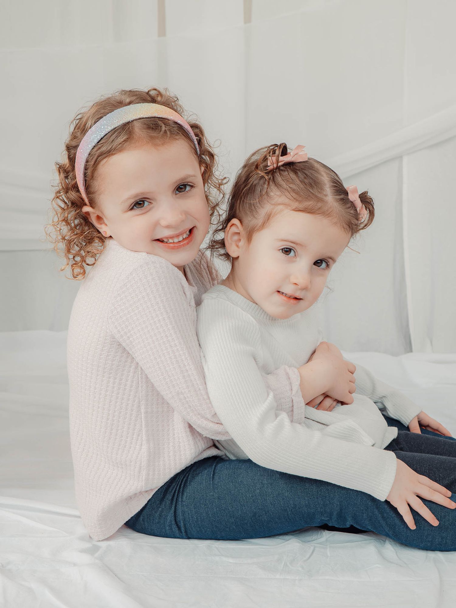 Two young sisters with curly hair sit together on a white bed, smiling warmly at the camera.