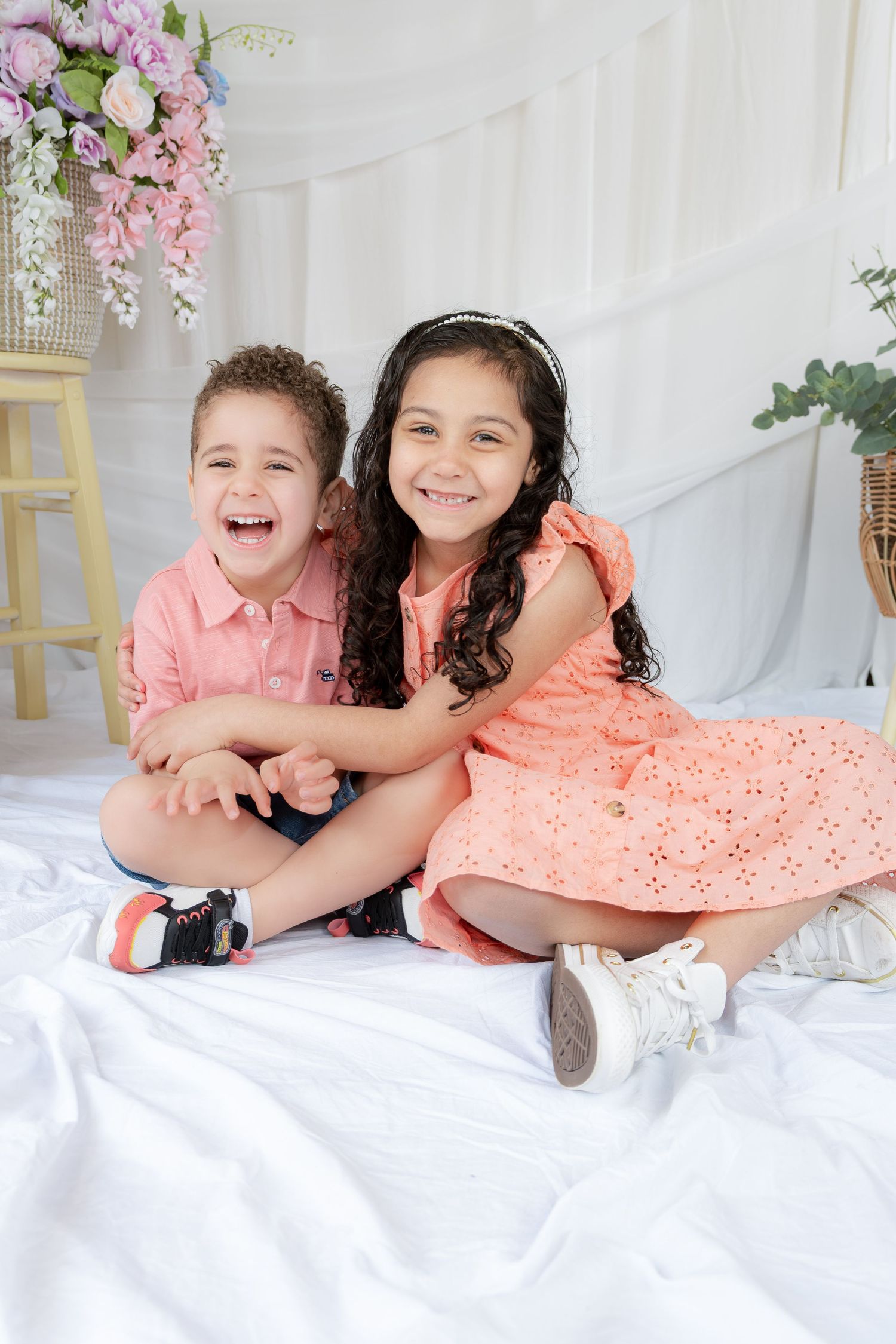 Two happy children in matching coral outfits sit together smiling on a white backdrop with flowers.