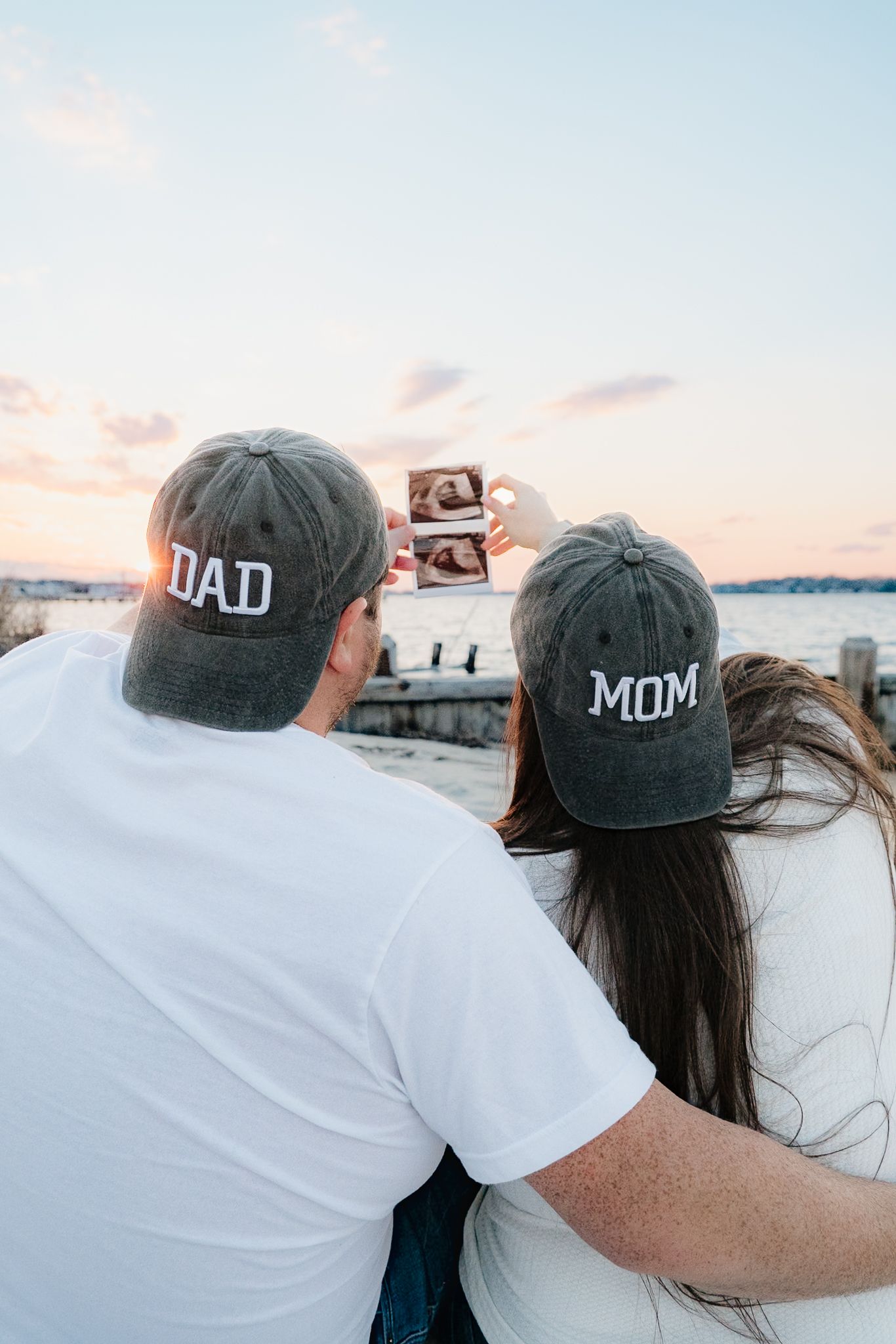 Couple wearing Dad and Mom hats holding ultrasound photos at sunset by the water, announcing pregnancy.