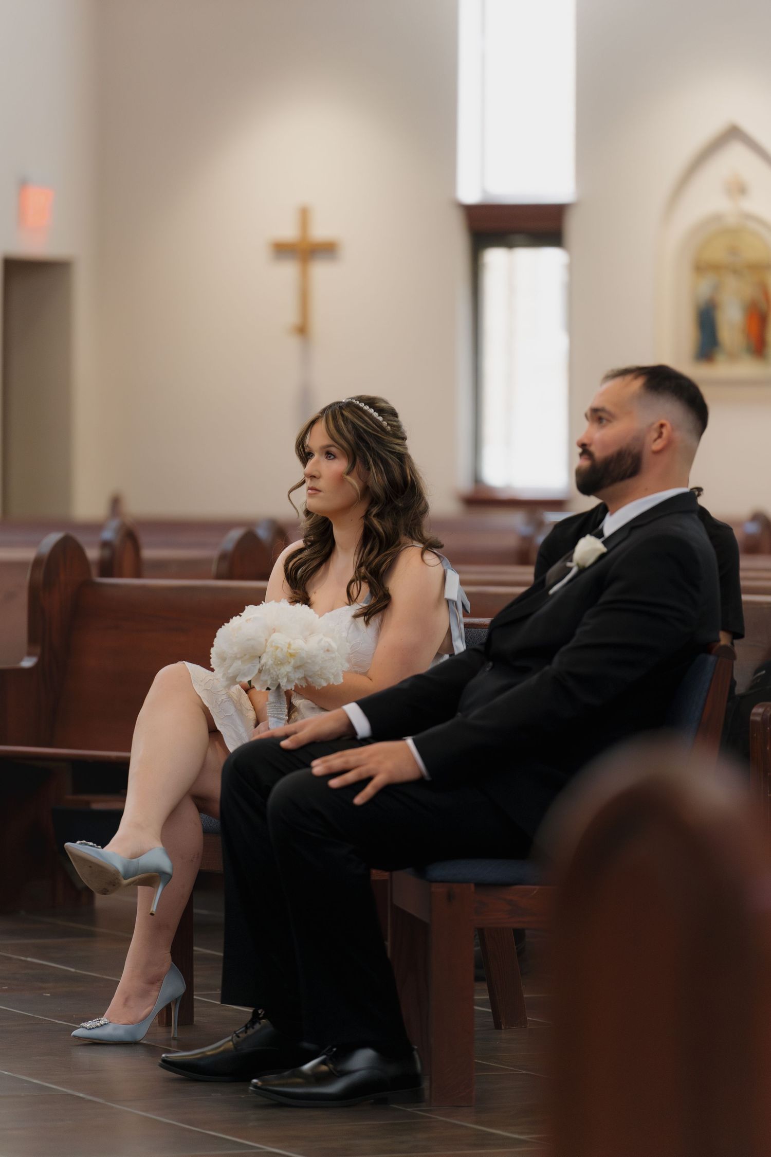 Bride and groom seated in church pews during wedding ceremony, bride holding white bouquet wearing blue heels.