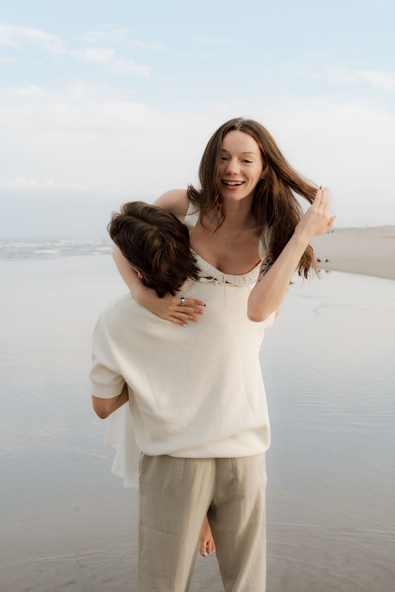 A joyful woman in a white dress gets a piggyback ride on the beach, laughing as waves roll in behind them.