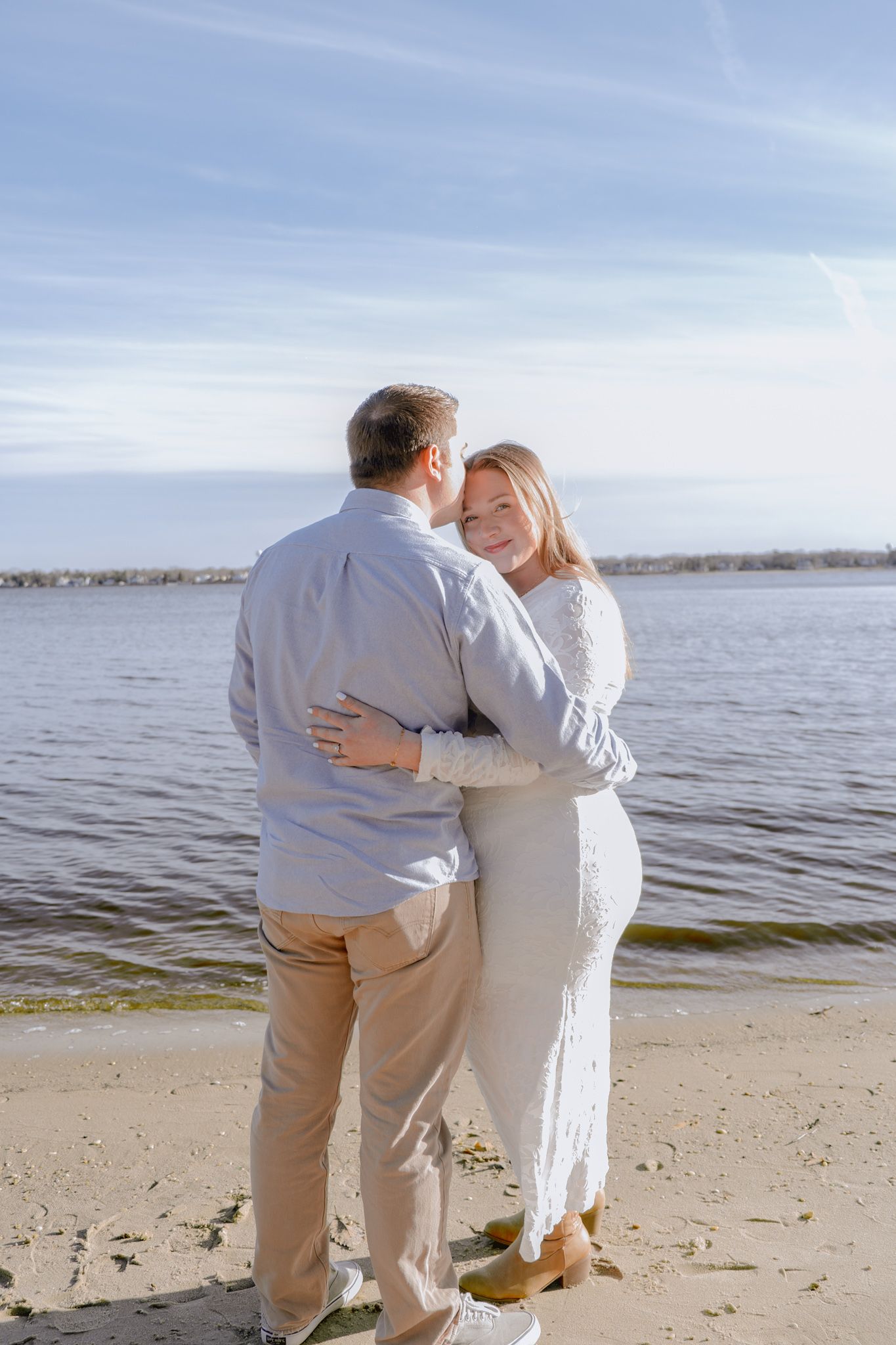 Engaged couple sharing a warm embrace on a sandy beach with sparkling water behind them.