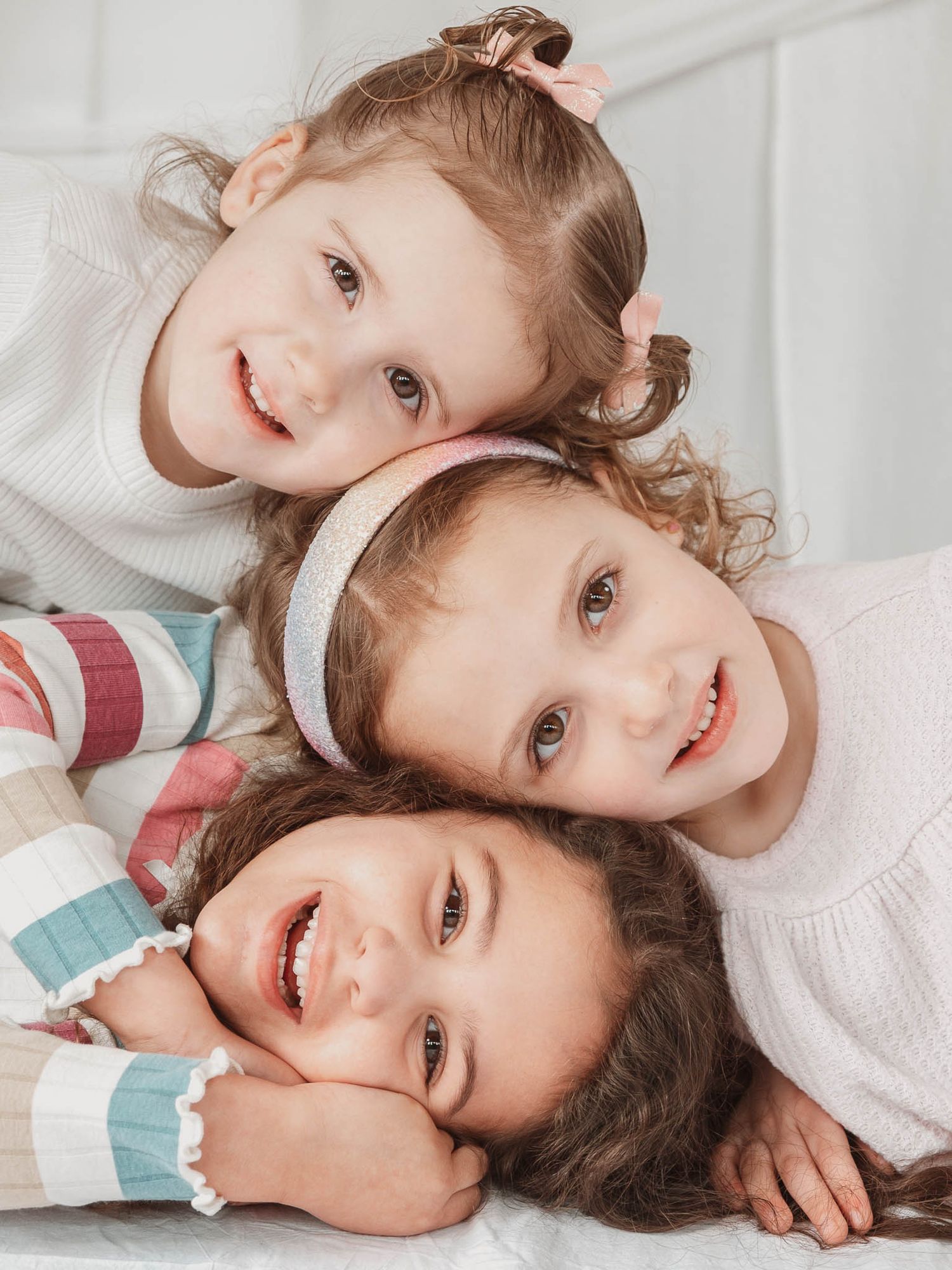 Three young girls with curly hair smile and pose together lying down in a bright white studio.