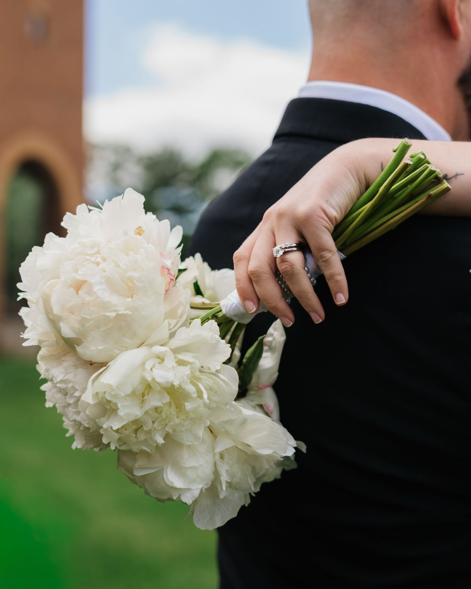 Bride holds white peony bouquet over grooms shoulder showing diamond engagement ring outdoors.