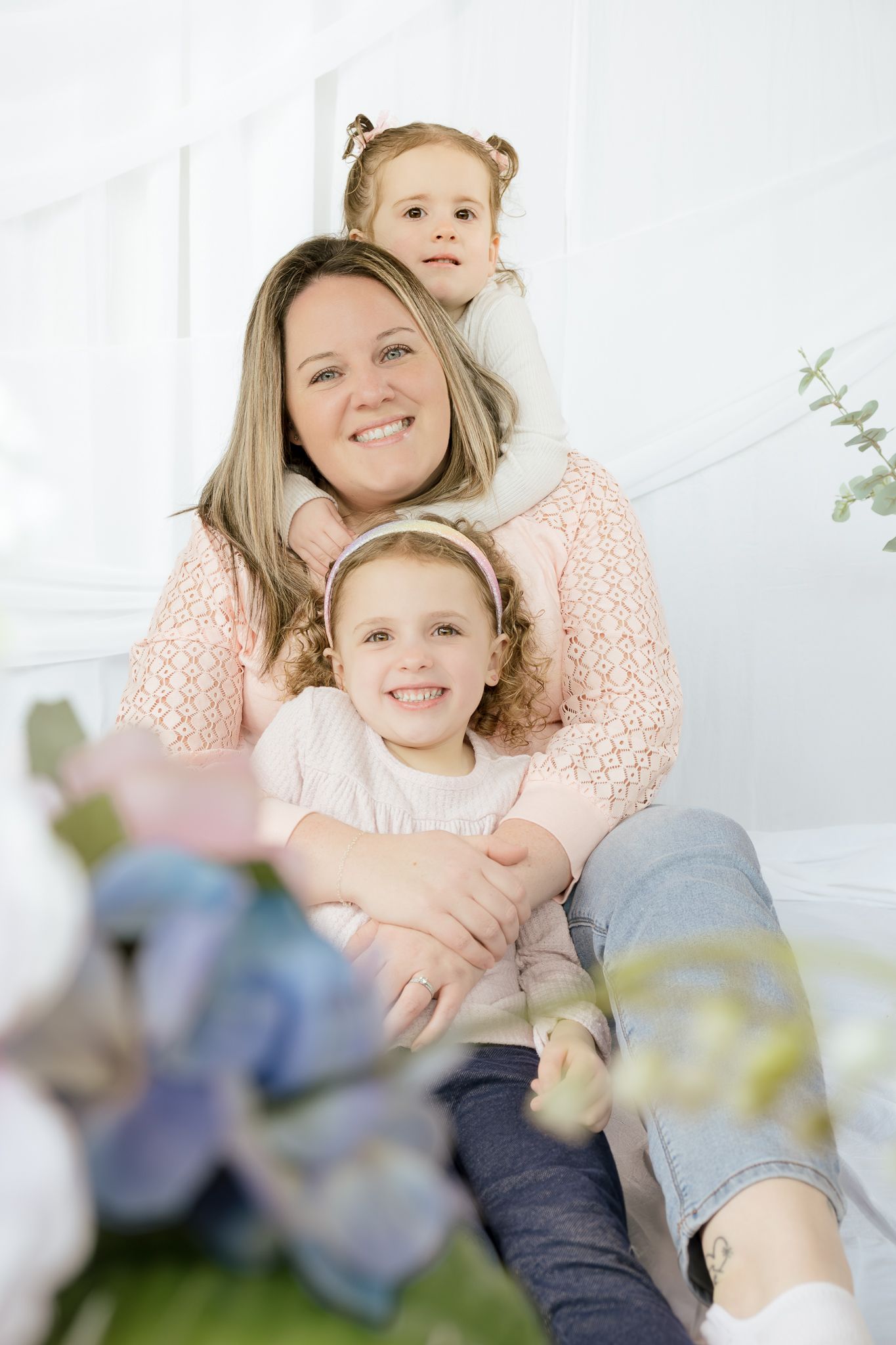 A smiling mother embraces her two young daughters in a bright white studio with floral decorations.