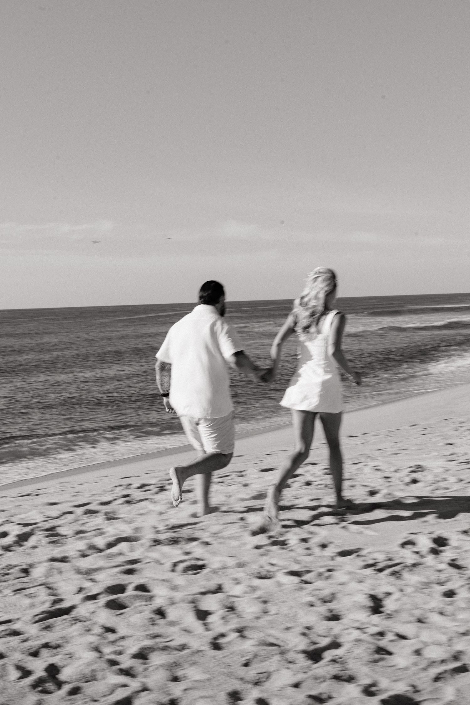 Black and white photo of a couple holding hands running barefoot on a sandy beach toward the ocean.