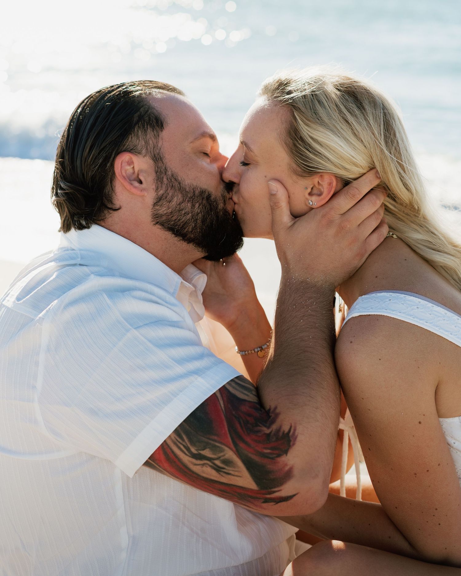 A couple shares a romantic kiss on the beach, man with beard and tattoo, woman in white top, ocean backdrop.