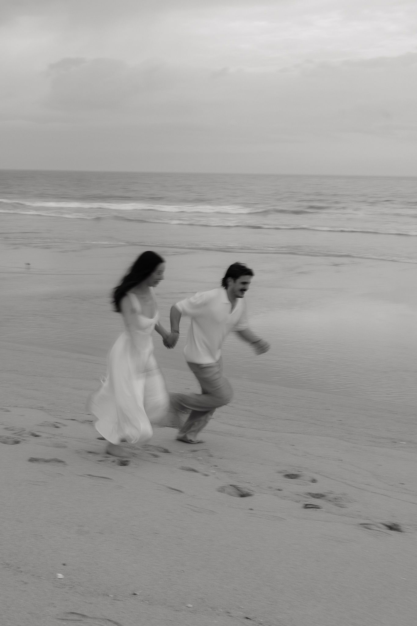 Black and white photo of a joyful couple running hand in hand on a sandy beach with ocean waves behind them.