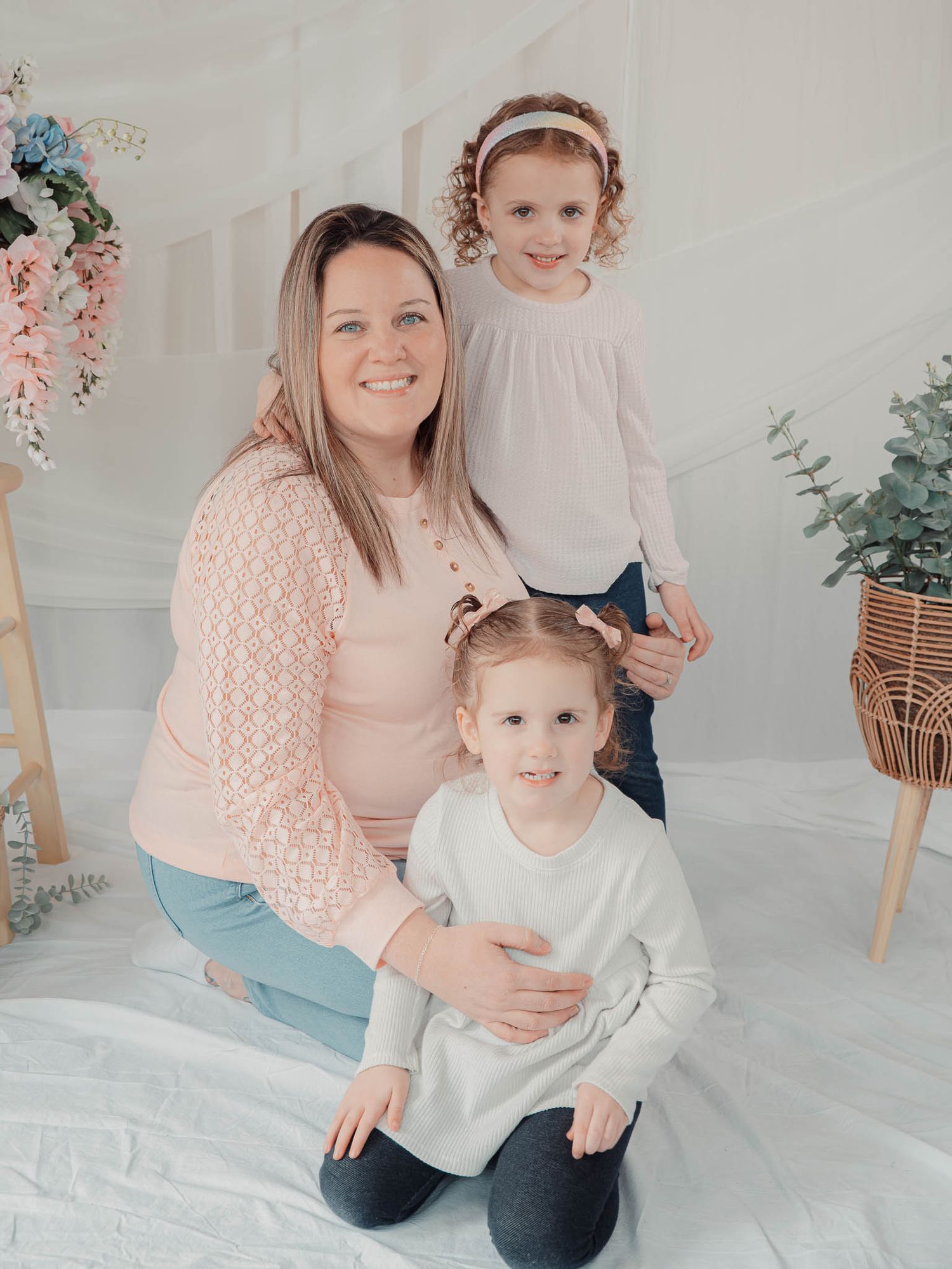 A smiling mother kneels with her two young daughters in a bright floral studio setting.