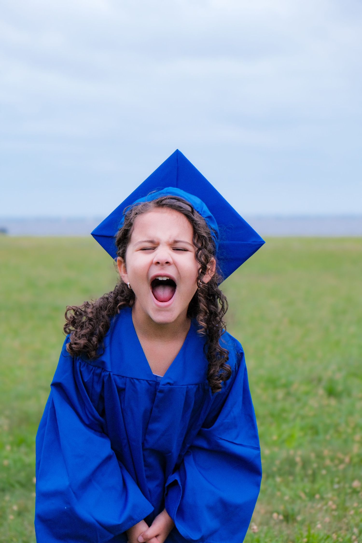 Young girl in blue graduation cap and gown shouting joyfully in an open green field.