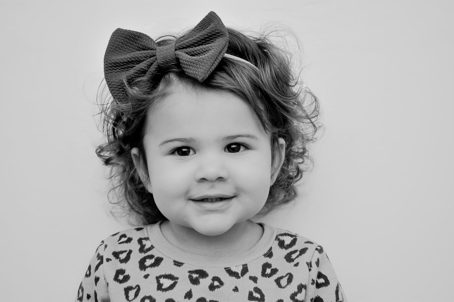 Black and white portrait of a cute toddler girl with curly hair and a bow headband.