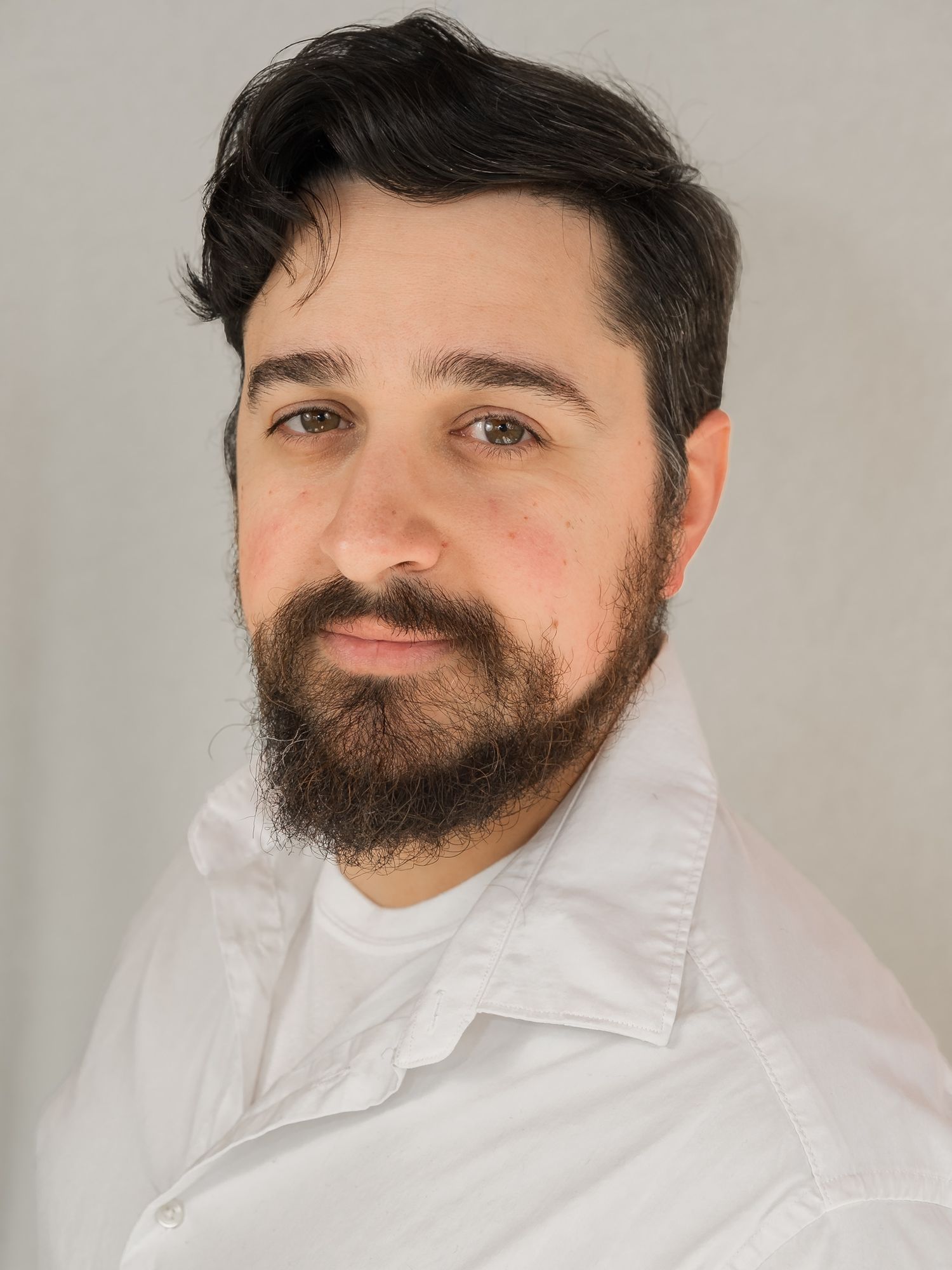 Bearded man with dark hair wearing a white shirt poses for a professional headshot against a gray background.