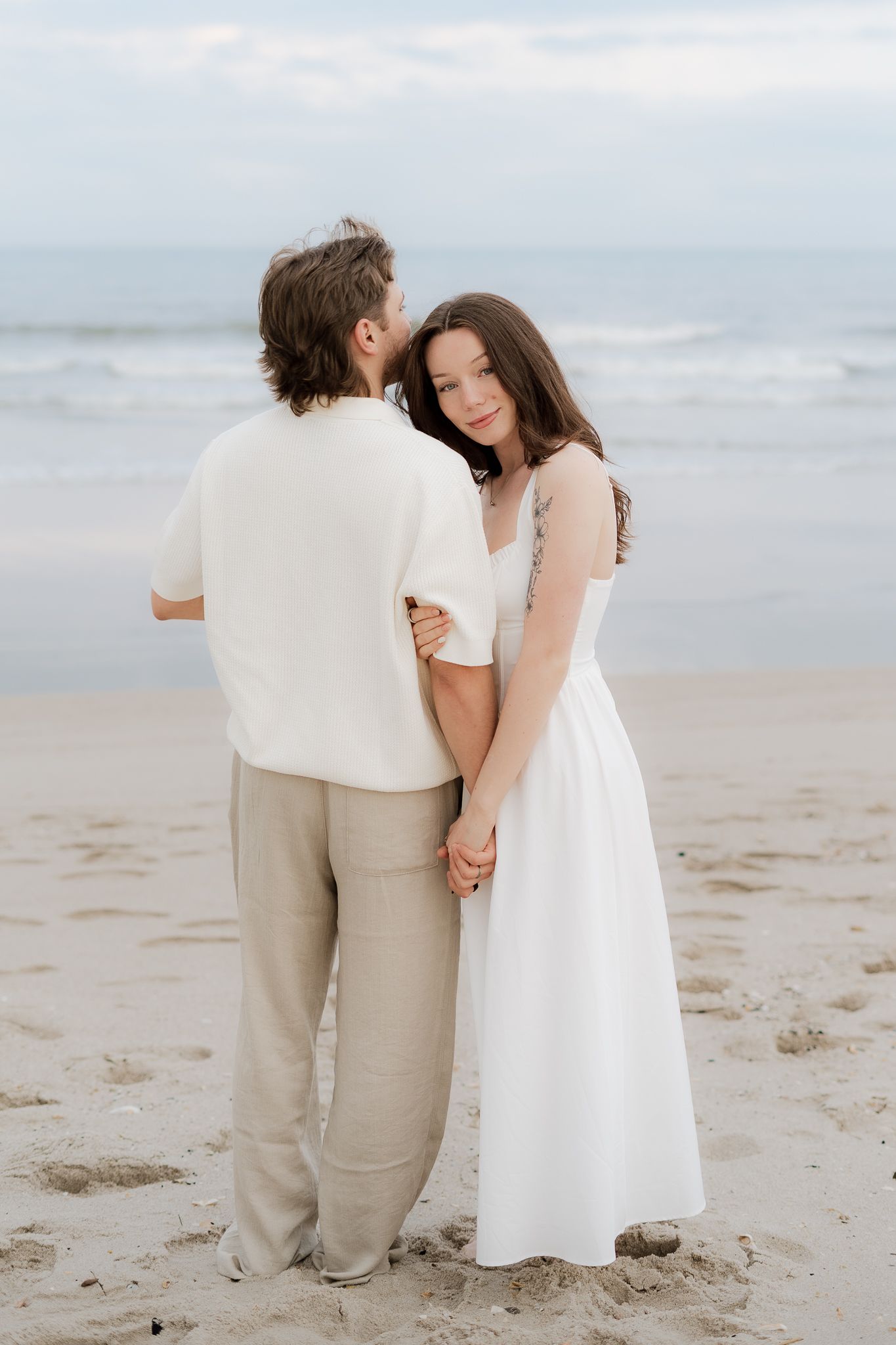 Couple holding hands on sandy beach, woman in white dress smiling softly, man in white shirt and tan pants facing ocean.